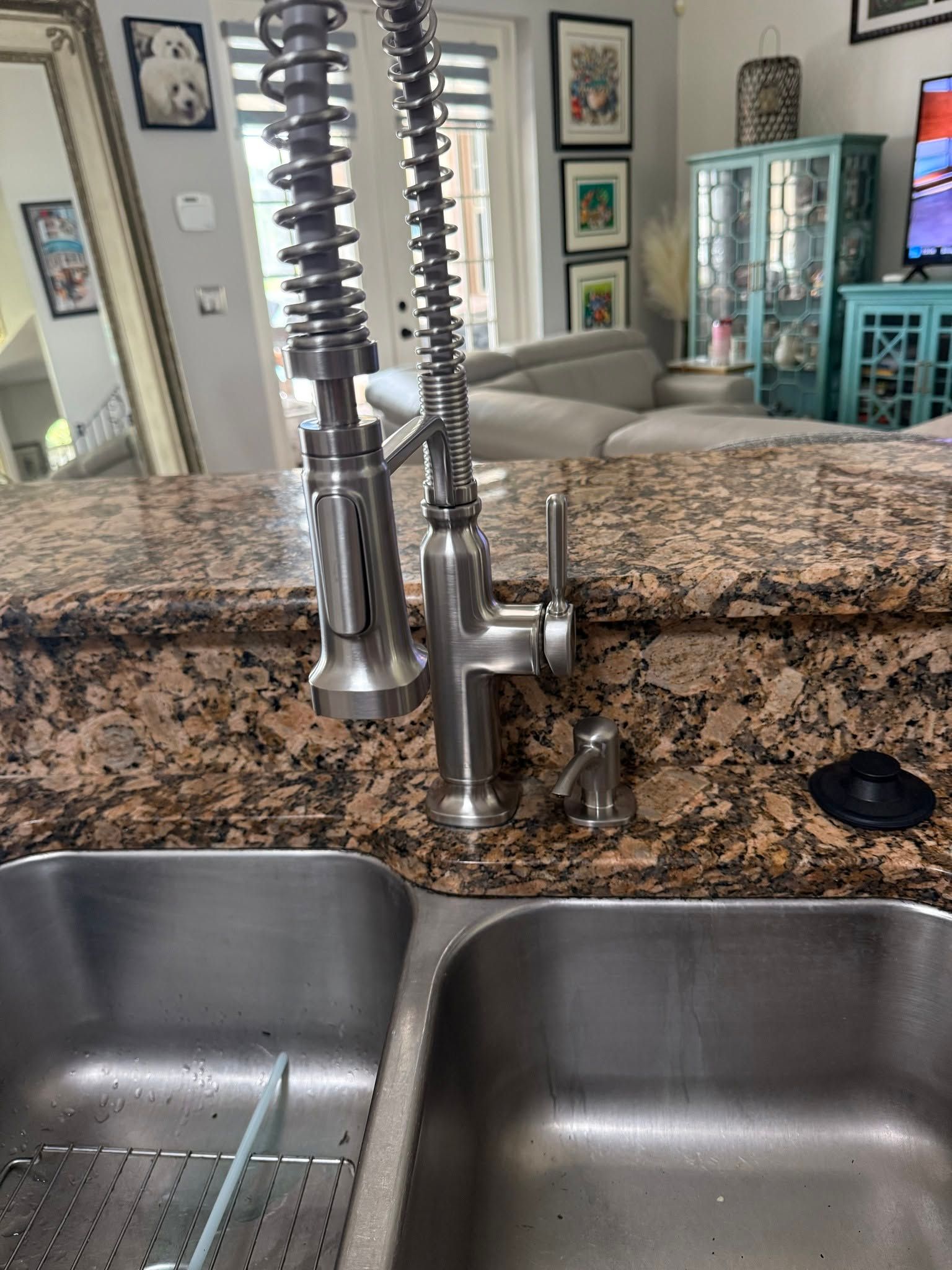Granite kitchen countertop with double stainless-steel sink and tall chrome faucet, reflecting a living room beyond