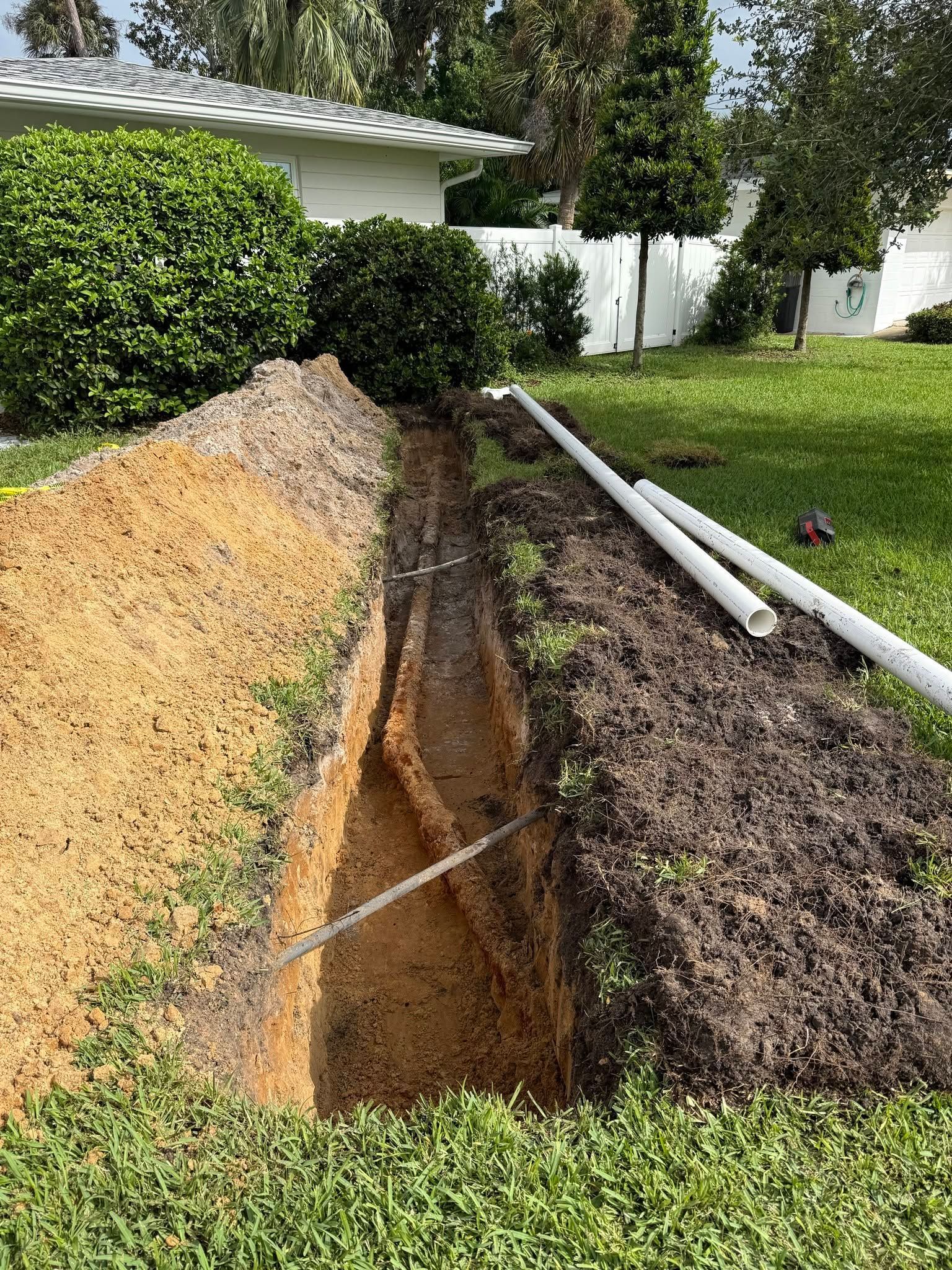 A deep trench dug beside a house, with dirt piles and white pipes along a grassy yard.