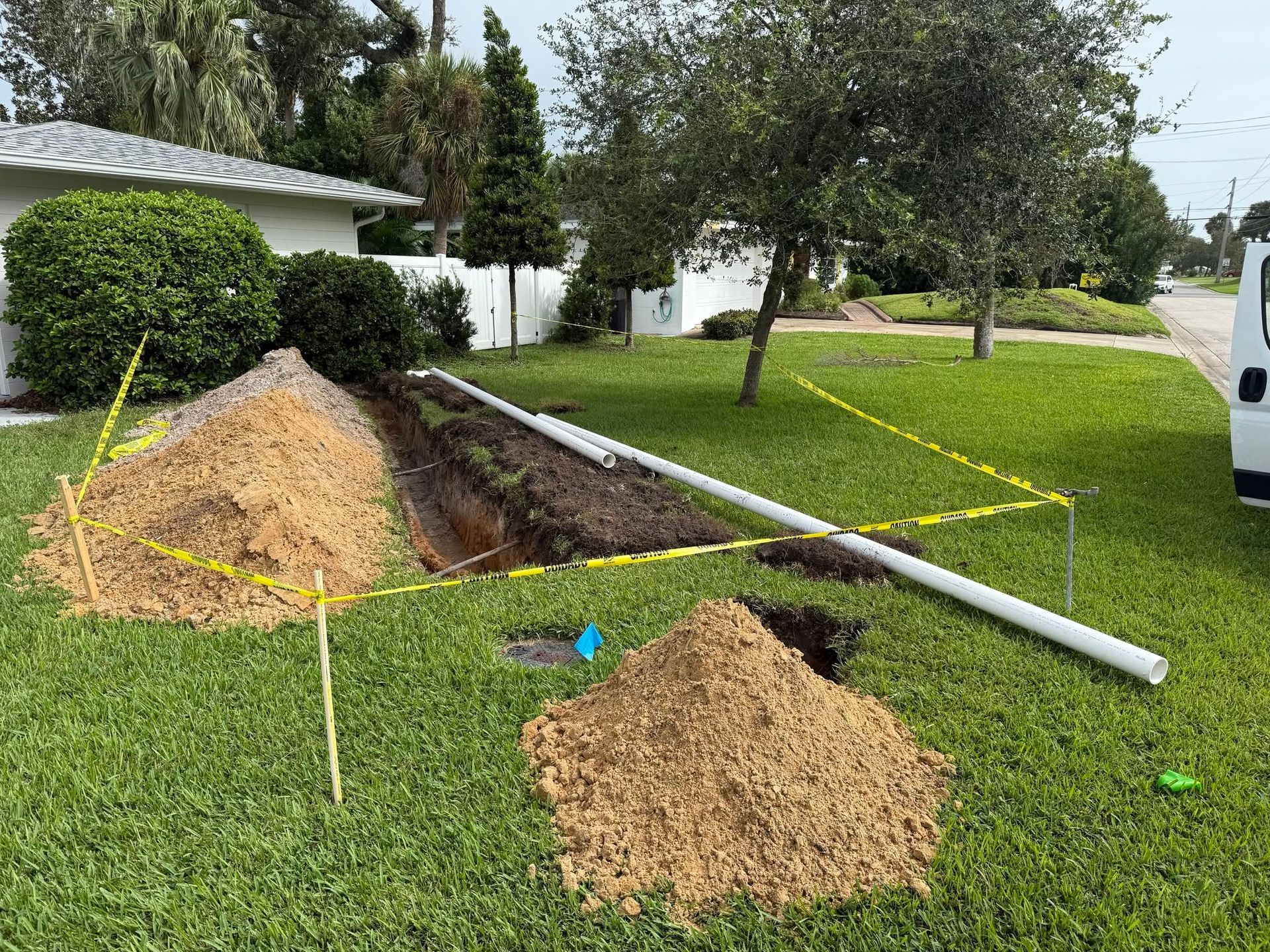 Utility trench with exposed pipe and two dirt piles in a grassy roadside yard, marked by yellow tape.