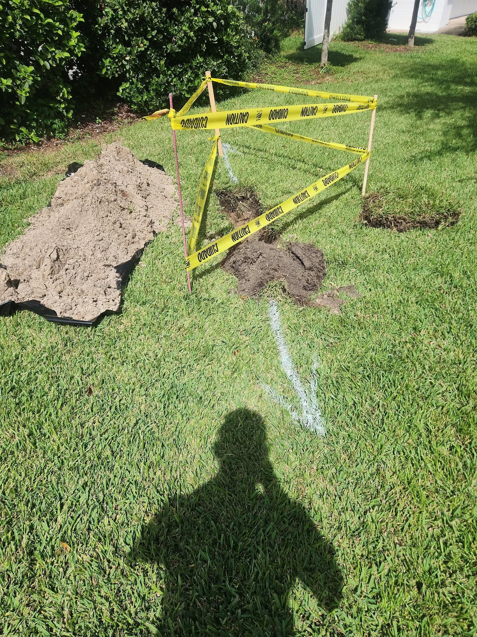 Caution tape around a disturbed lawn with dirt piles and a chalk marking near a buried utility line.