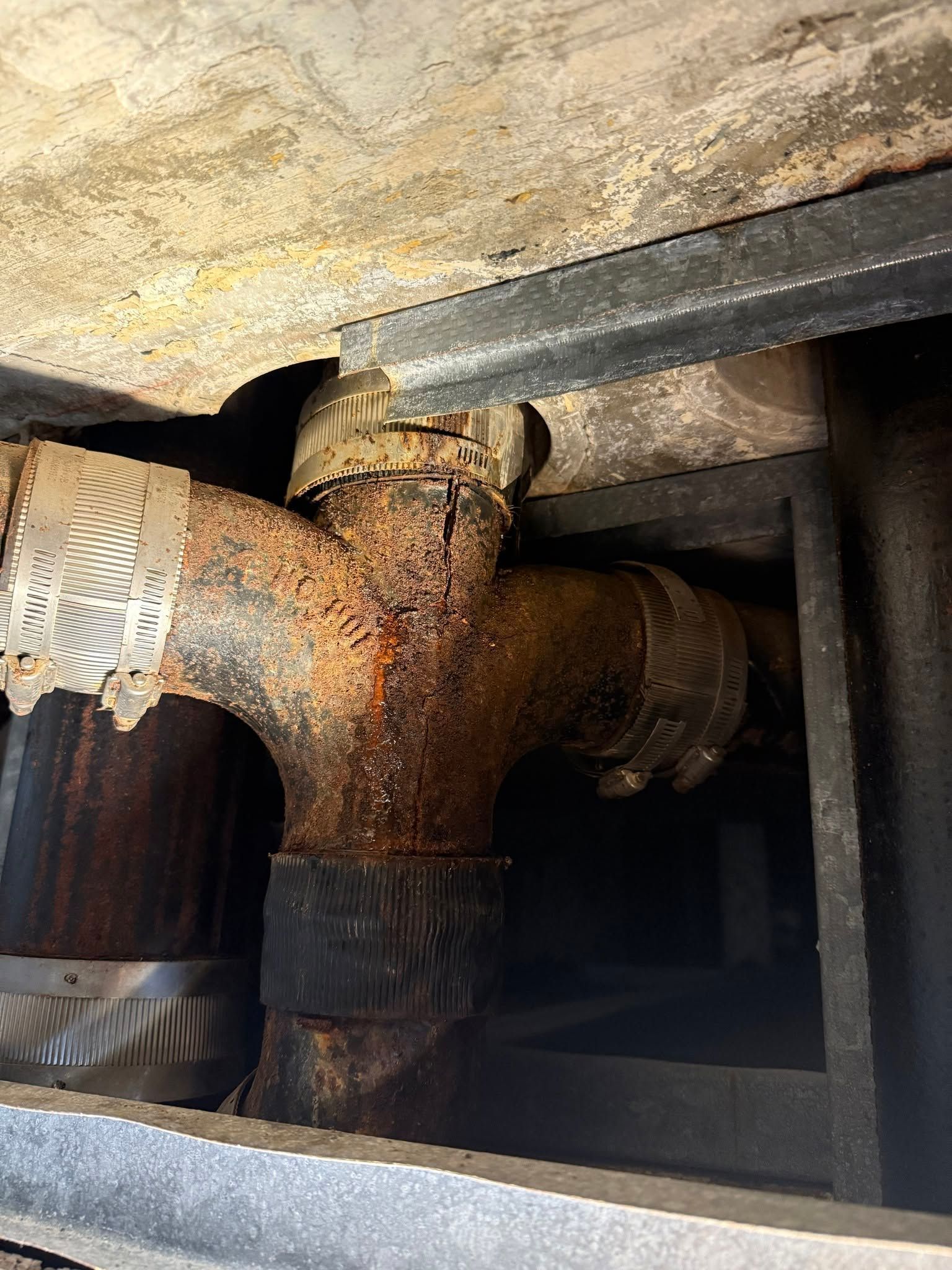 Rusty pipe junction under a floor, with corroded metal and dark support beams.