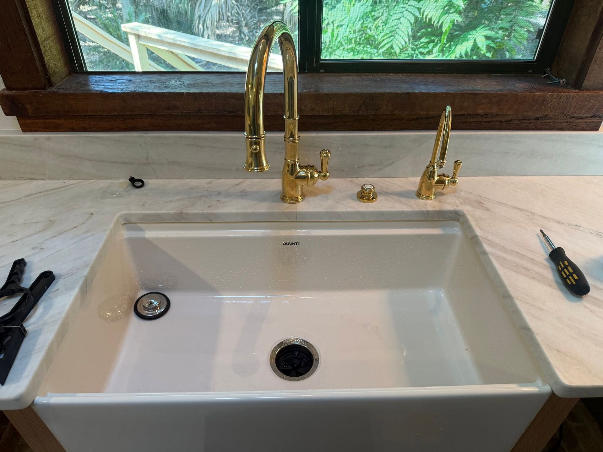 White farmhouse sink with gold faucet and soap dispenser on a marble countertop by a window