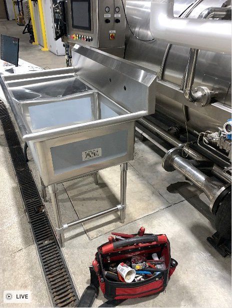 Stainless-steel industrial sink beside a large machine, with a red tool bag on the floor in a factory setting