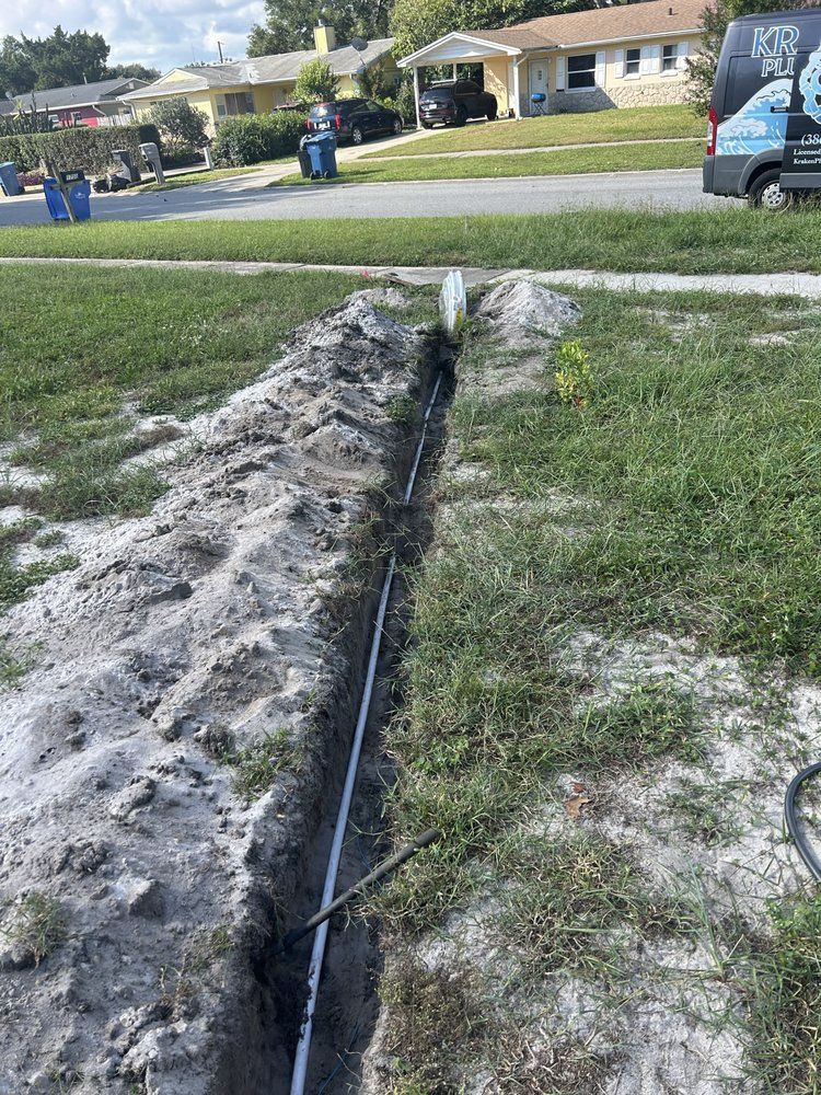 Trench cut through a grassy yard with a black pipe laid in it, near houses and workers in the background