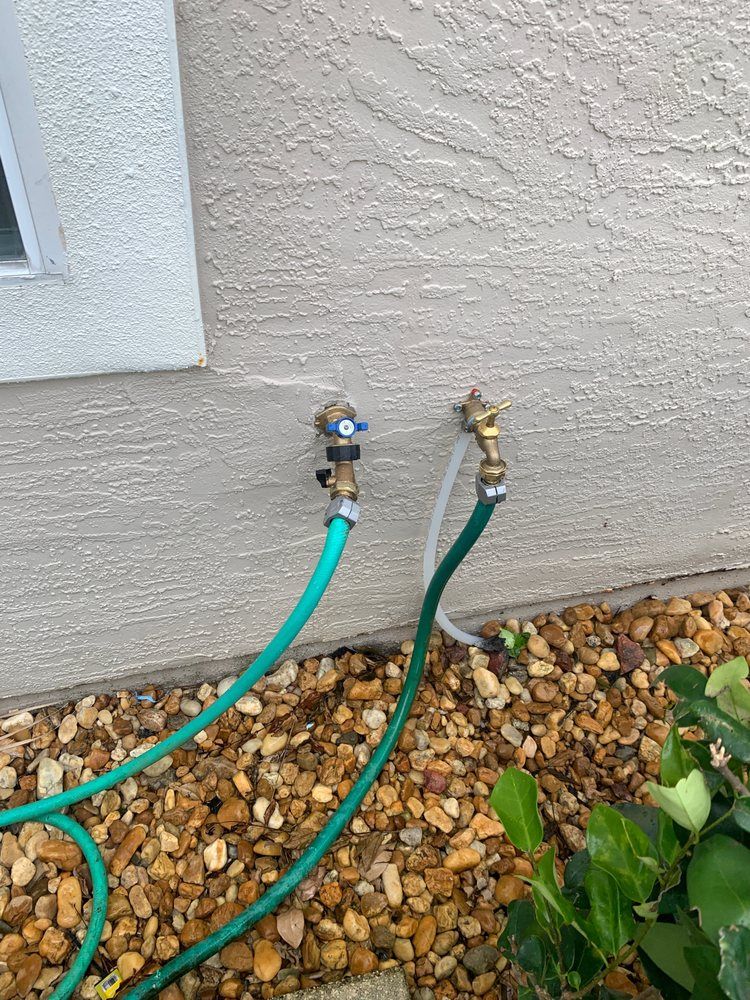 Two outdoor hose bibs on a stucco wall, each with a green garden hose attached.