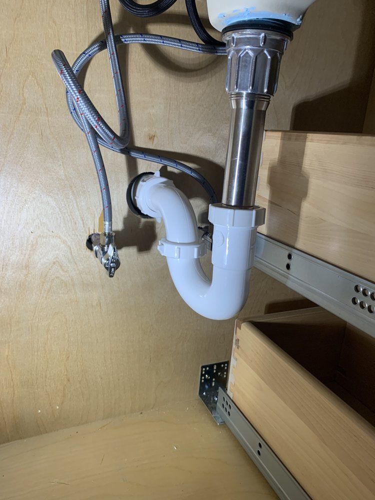 Under-sink plumbing with white drain trap and chrome pipe inside a wooden cabinet