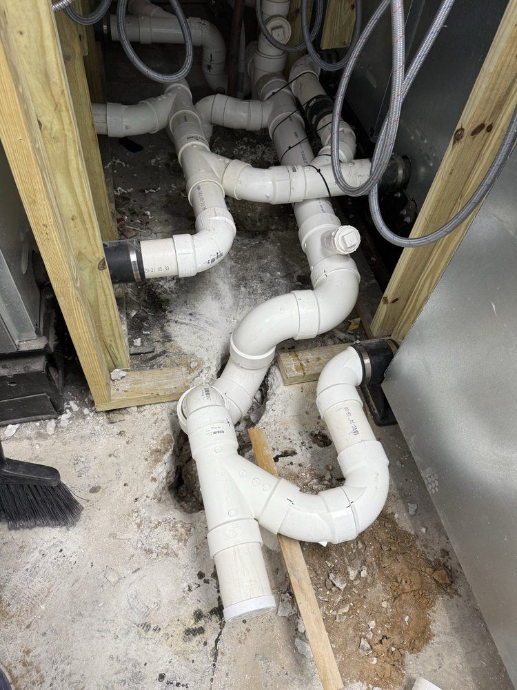 White PVC plumbing pipes and fittings in a cabinet opening on a dirty floor, with exposed walls and debris.