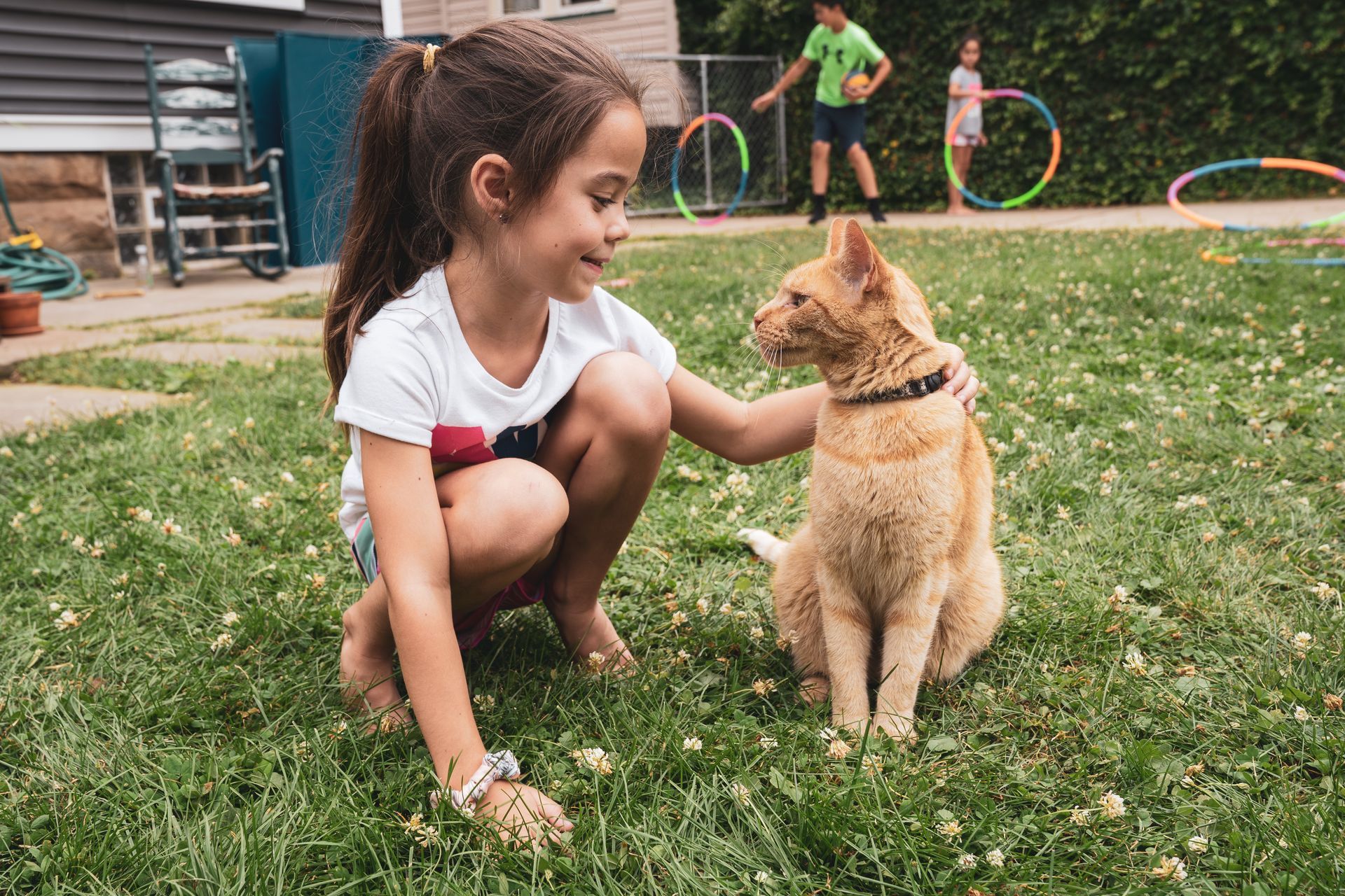 A little girl is petting an orange cat in the grass.