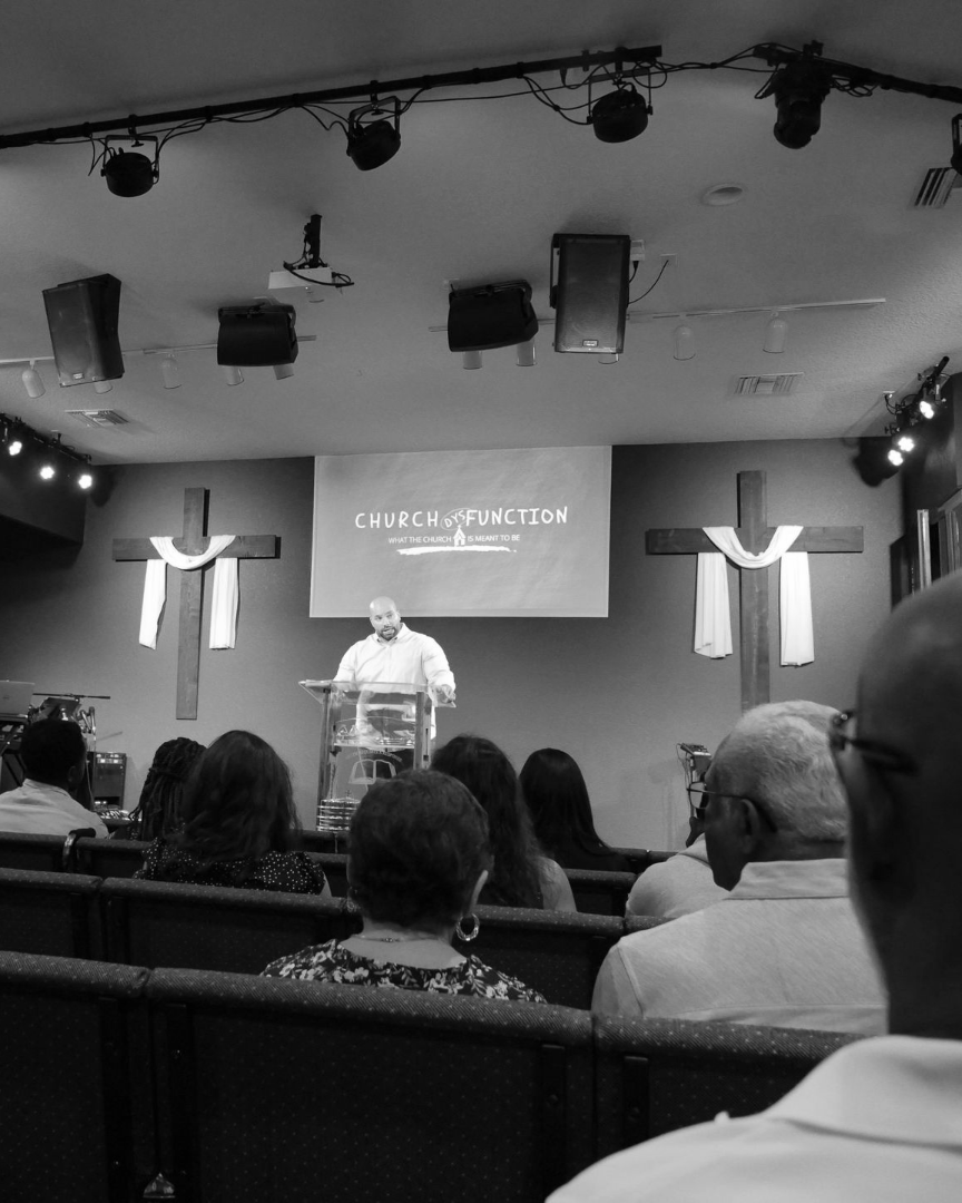 Person speaking at church podium with audience seated and cross symbols on the walls