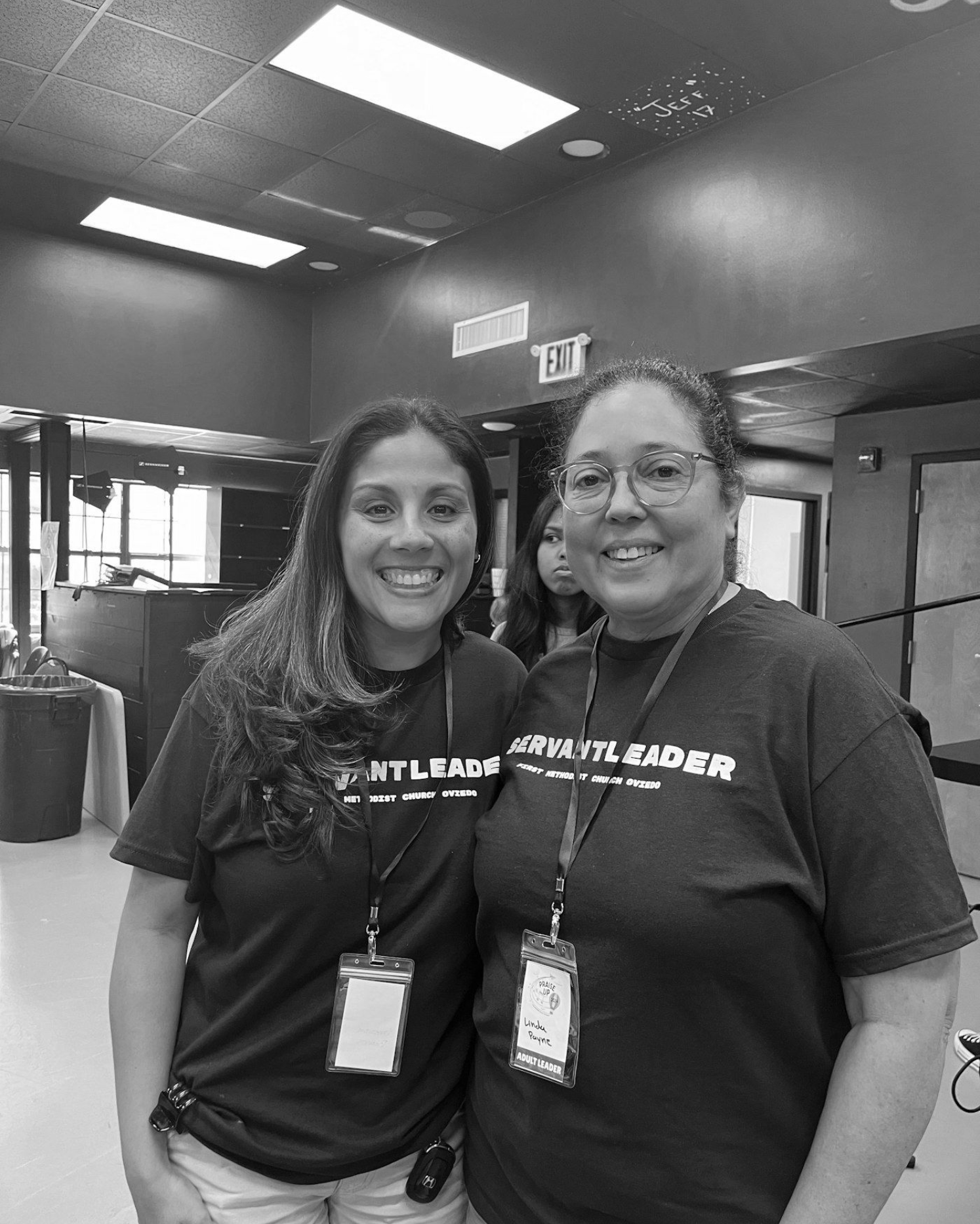Two employees smiling indoors under bright ceiling lights, wearing matching T-shirts and lanyards.