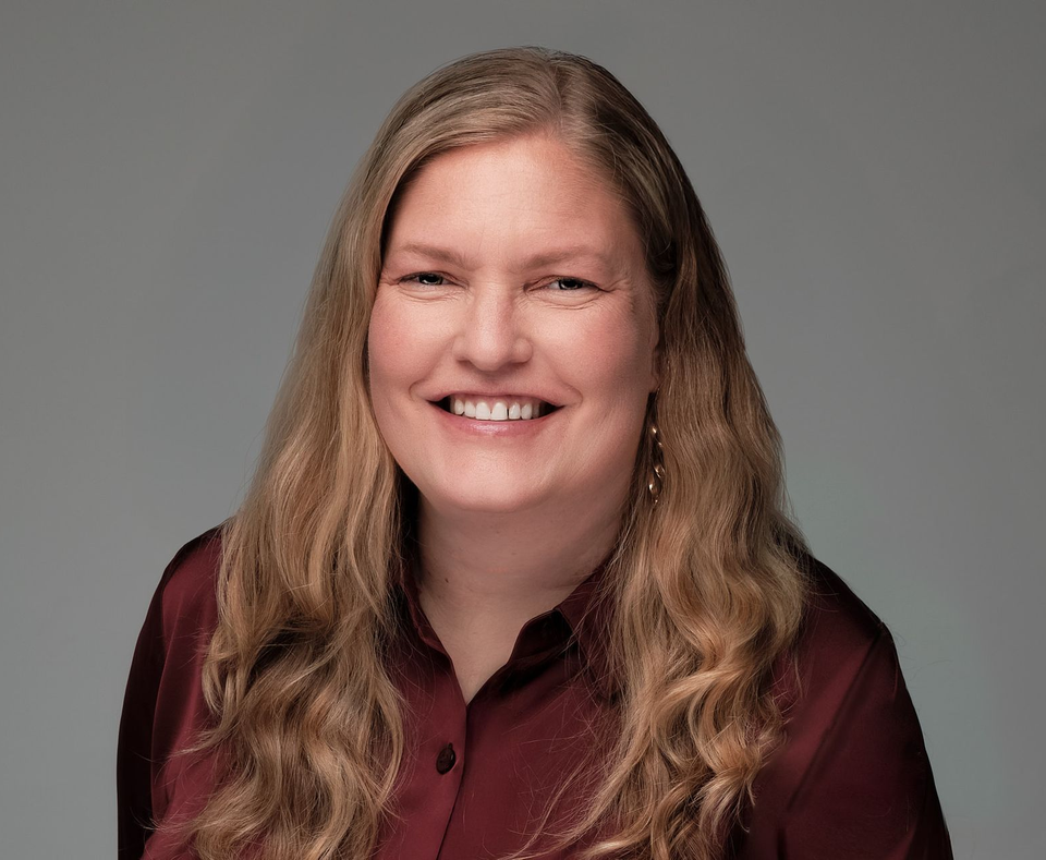 Smiling person with long, wavy hair wearing a maroon collared shirt against a solid grey background.