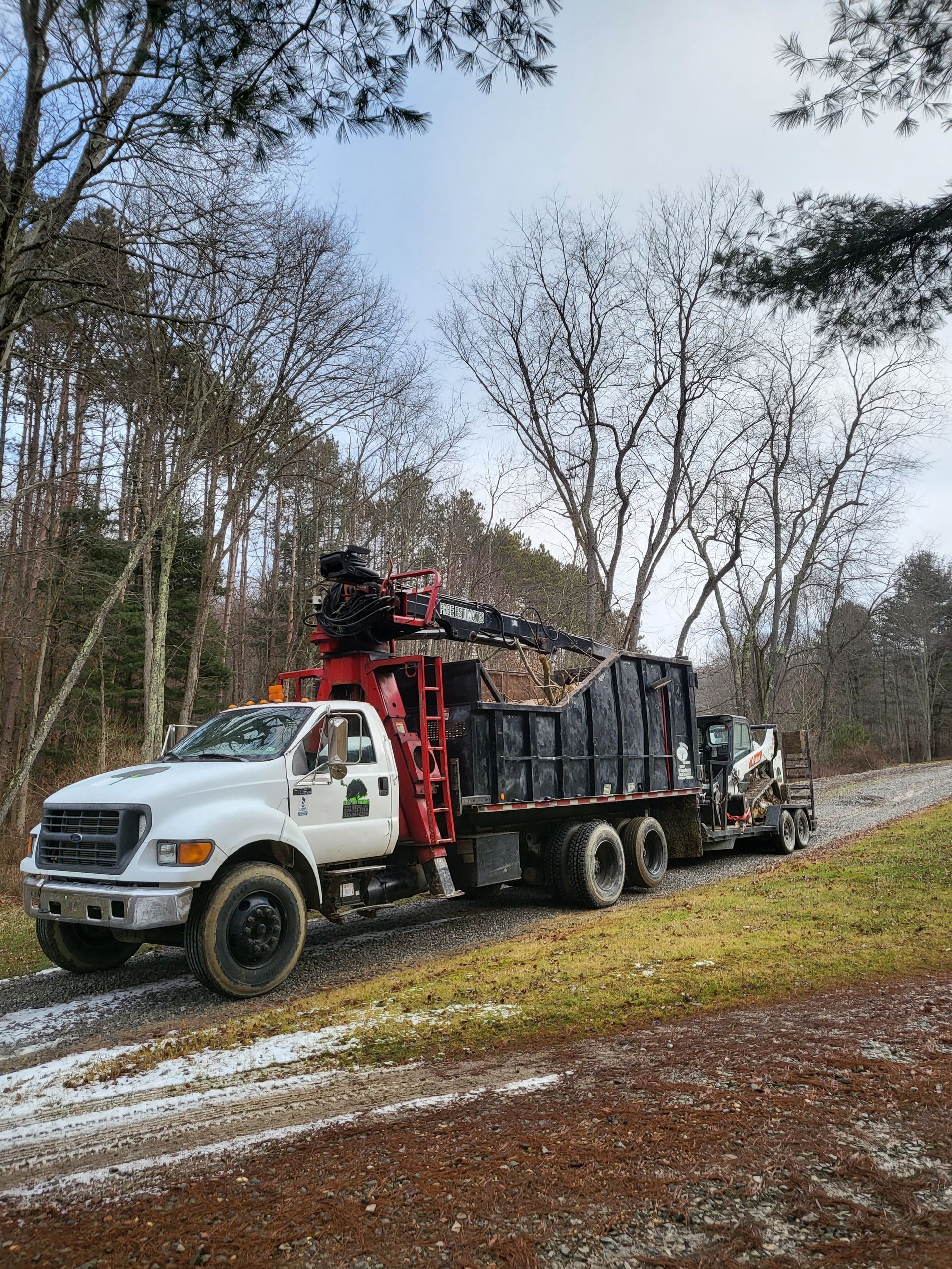 Truck carrying black-wrapped bales on a road, with a small excavator. Winter scene, leafless trees, overcast sky.
