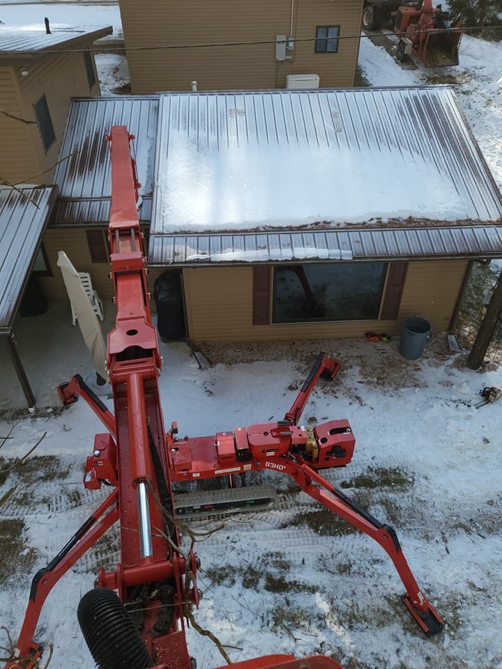 Red boom lift angled towards a snow-covered house roof. Winter scene with trees and other buildings.