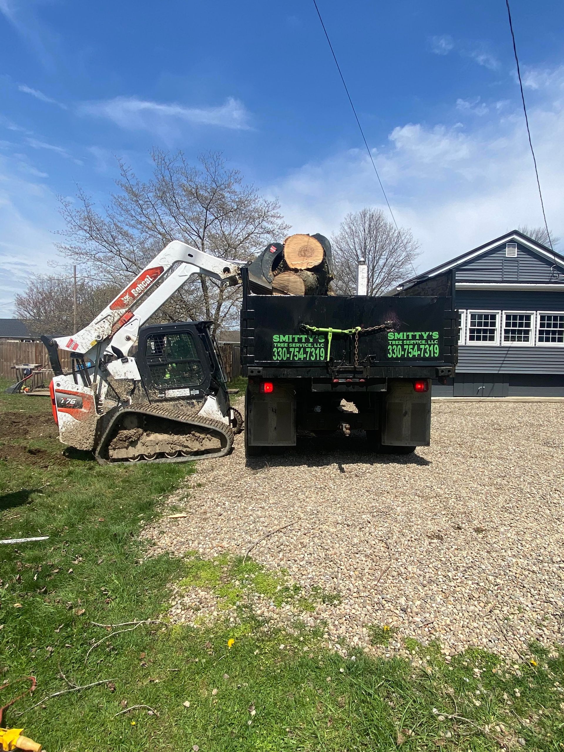 Bobcat loader loading logs into a black dump truck parked on gravel next to a house with a blue sky.
