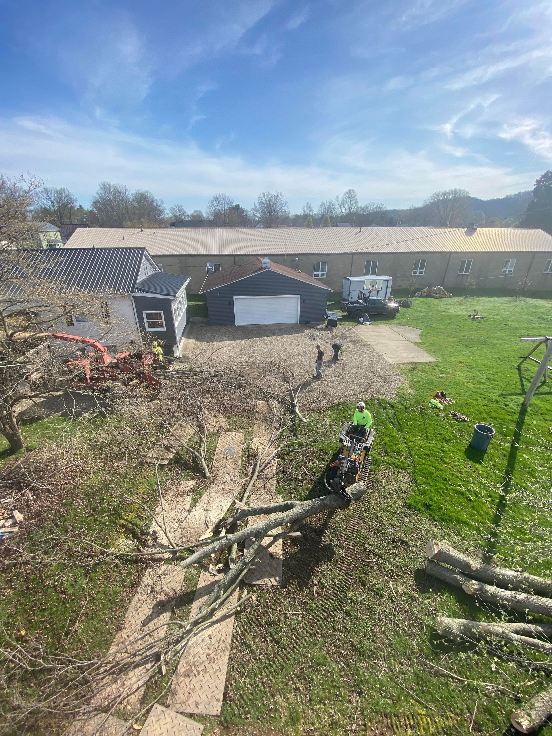 Yard work scene: Two people near a garage with a small shed, logs, and a green lawn under a partly cloudy sky.