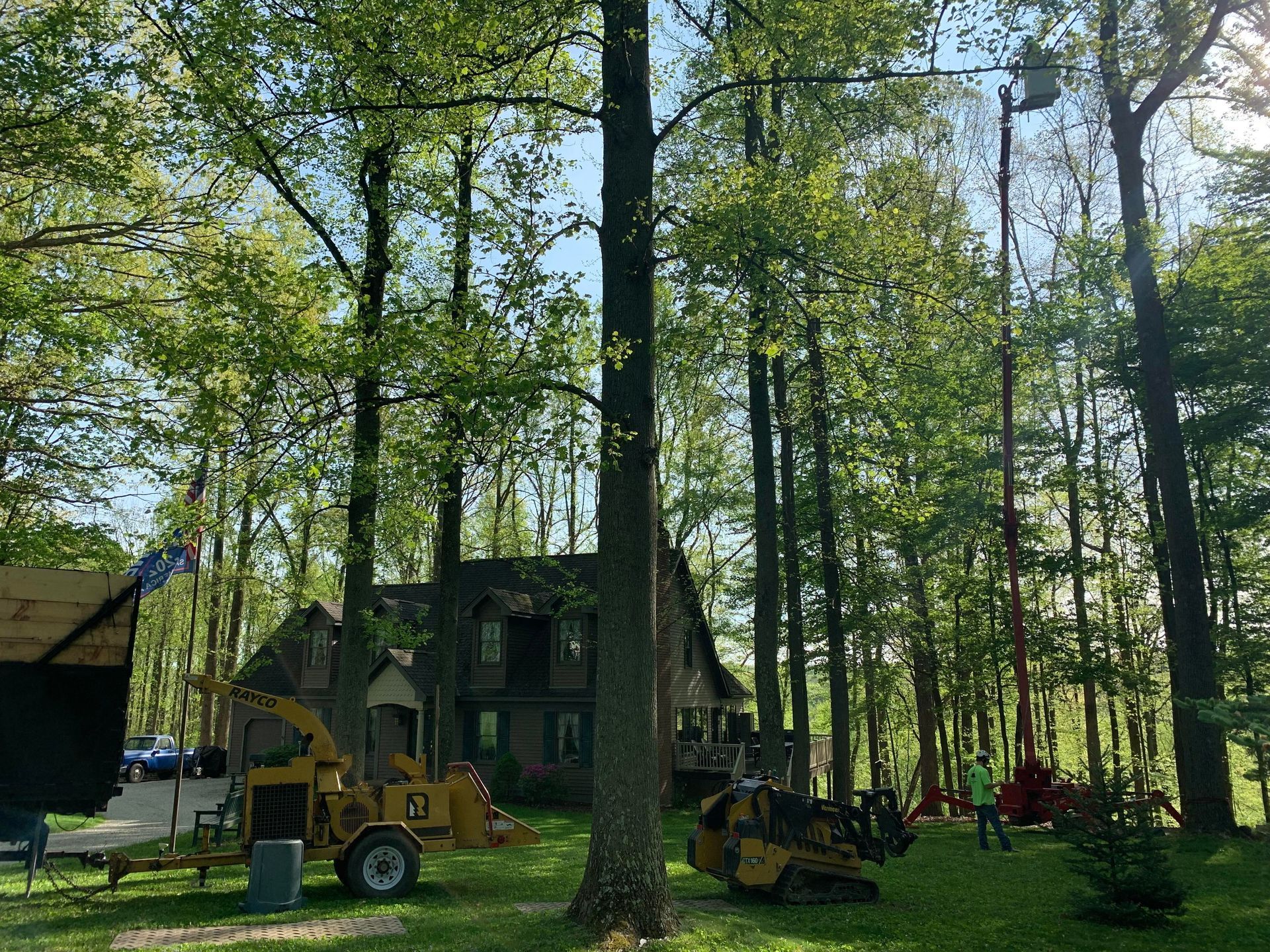 Tree removal in progress near a house; chipper, lift, and machinery present in a wooded setting.