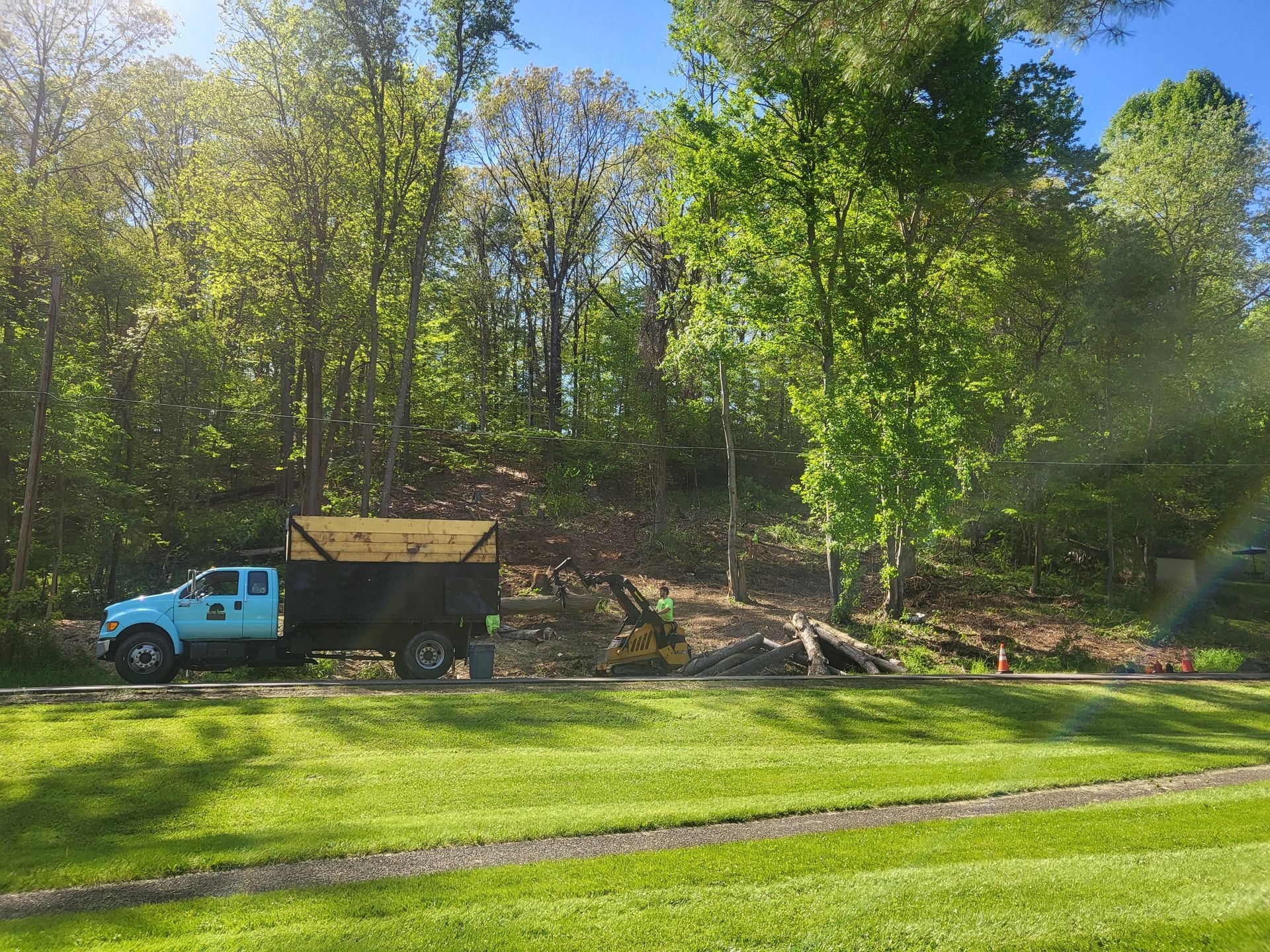 Blue truck parked, loading wood debris near wooded area on a sunny day.
