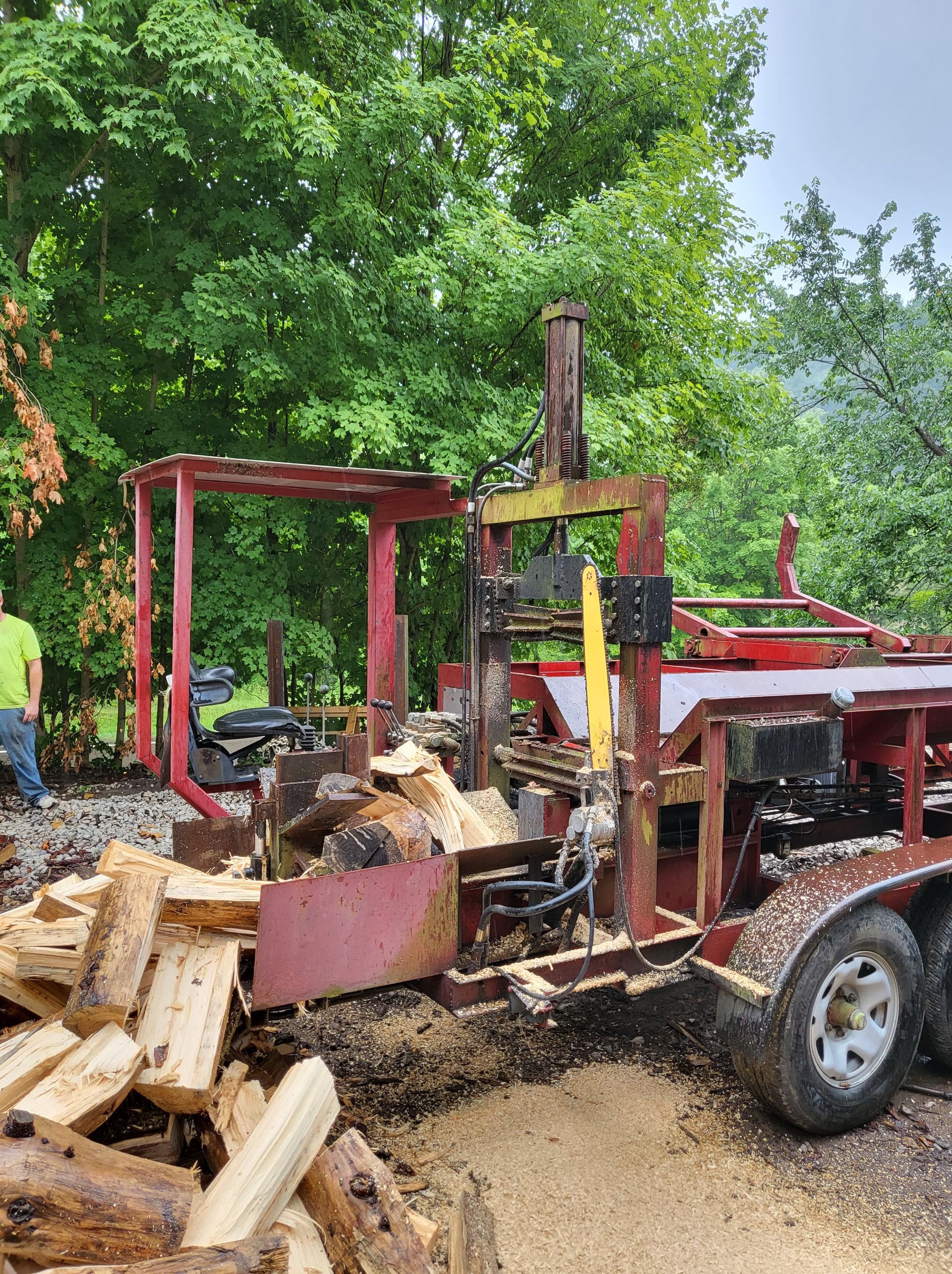 Wood splitter processing logs; red machine, pile of split wood, person nearby.