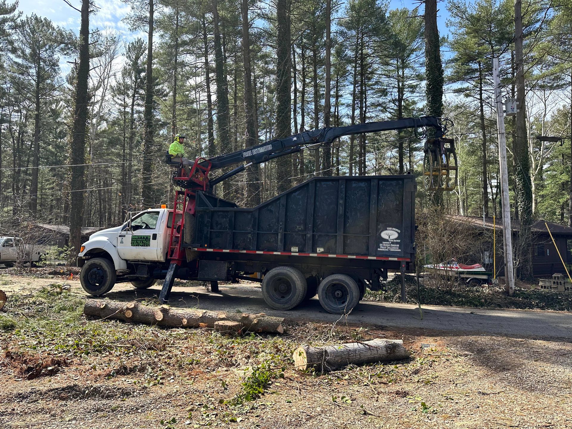 Truck with a crane, loading wood debris in a forest. A worker operates the crane.