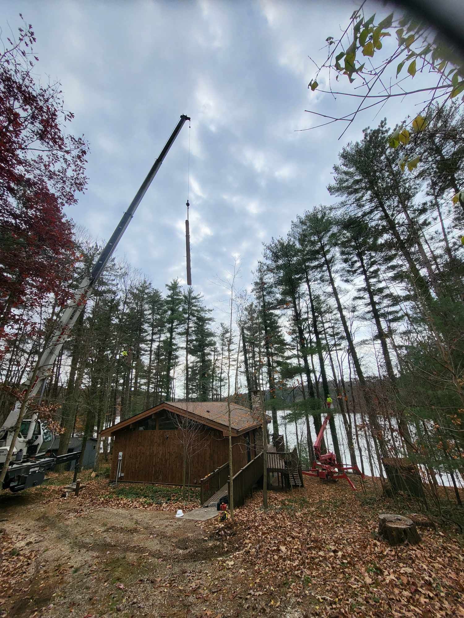 A crane lifts a tall structure over a cabin near a lake surrounded by trees. Cloudy sky.