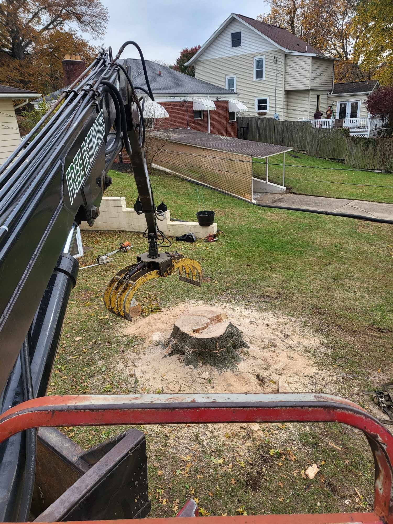 An excavator removing a tree stump in a yard with houses in the background.