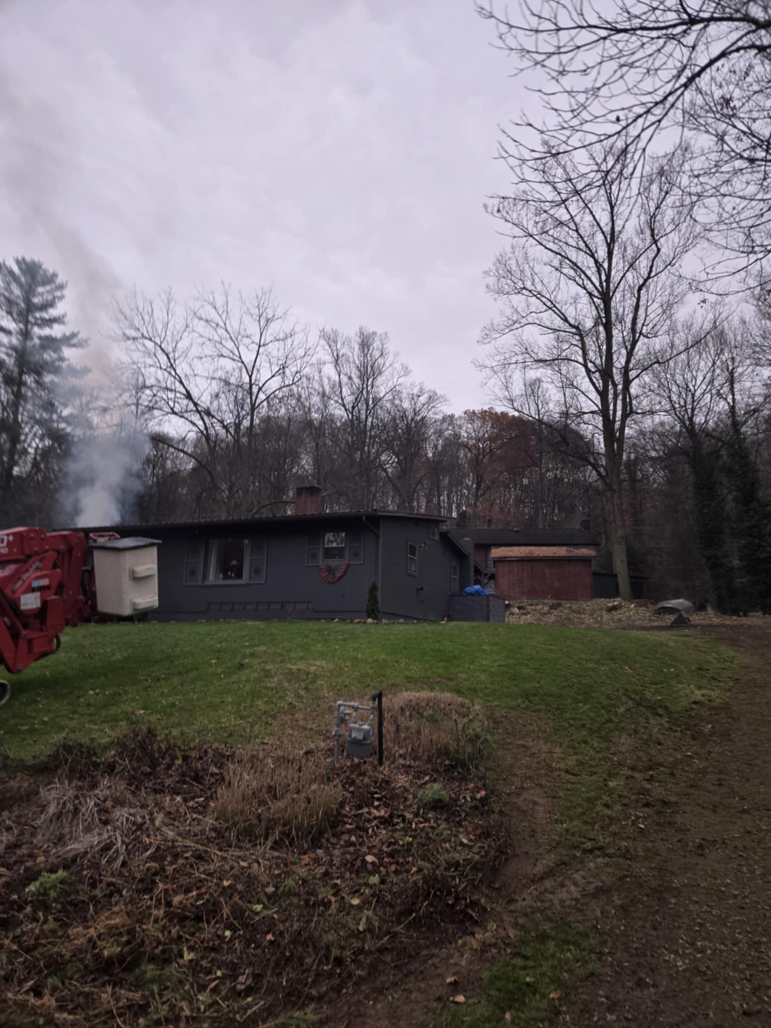 A one-story gray house with smoke rising from a chimney, in front of bare trees under an overcast sky.