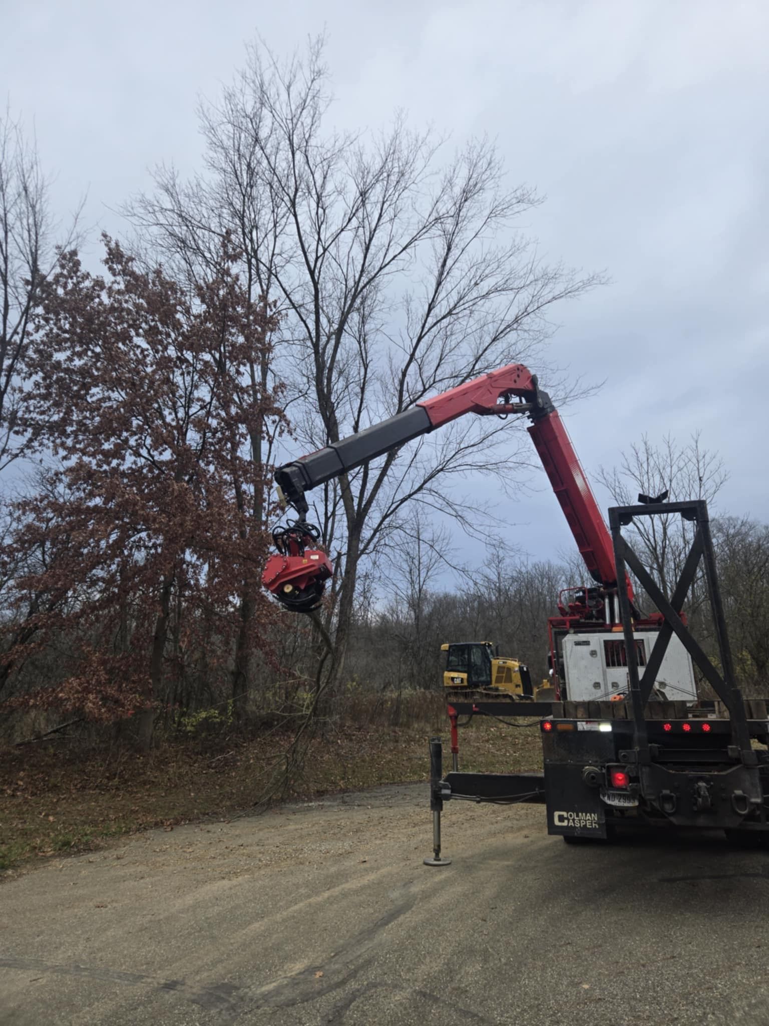 Red crane with a tree shear trimming a tree on a gravel road. A bulldozer is in the background.