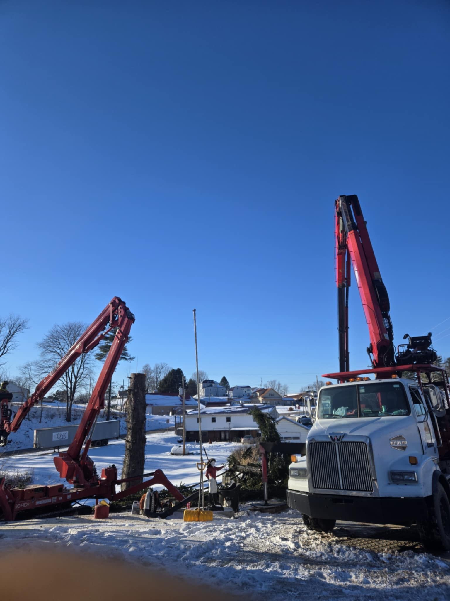 Two red boom trucks operating in a snowy area, with buildings and a flagpole in the background. Bright blue sky.