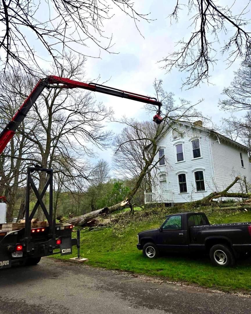A truck with a crane trimming a tree next to a white house with a black pickup truck parked nearby.