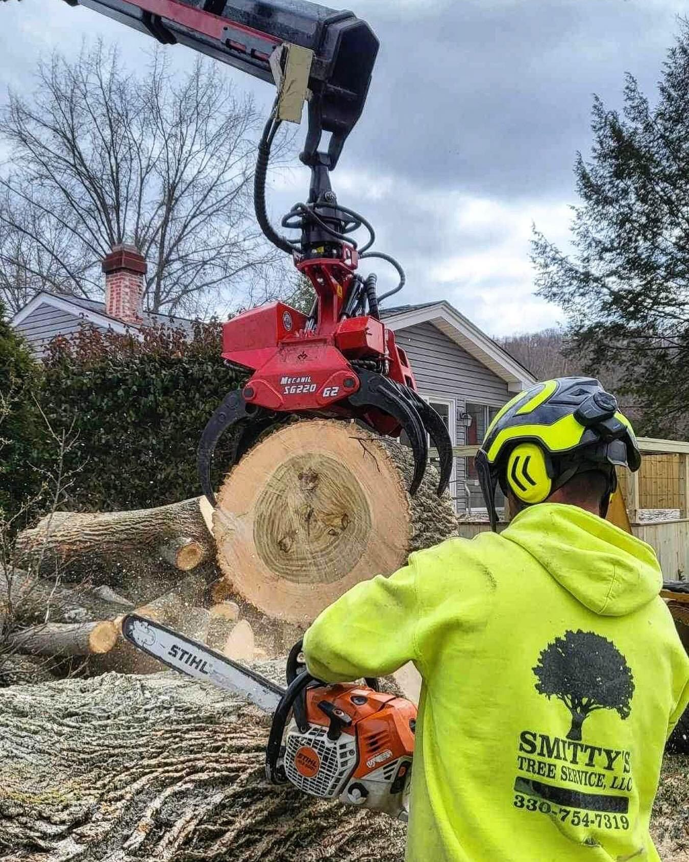 Worker using a chainsaw to cut a log held by a grapple. Bright yellow shirt, safety helmet. Outdoor setting.
