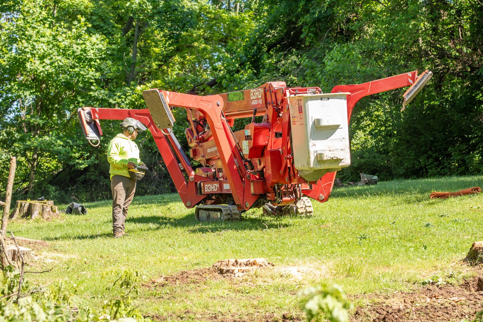 A worker in yellow vest operates a red stump grinder in a grassy field with trees.
