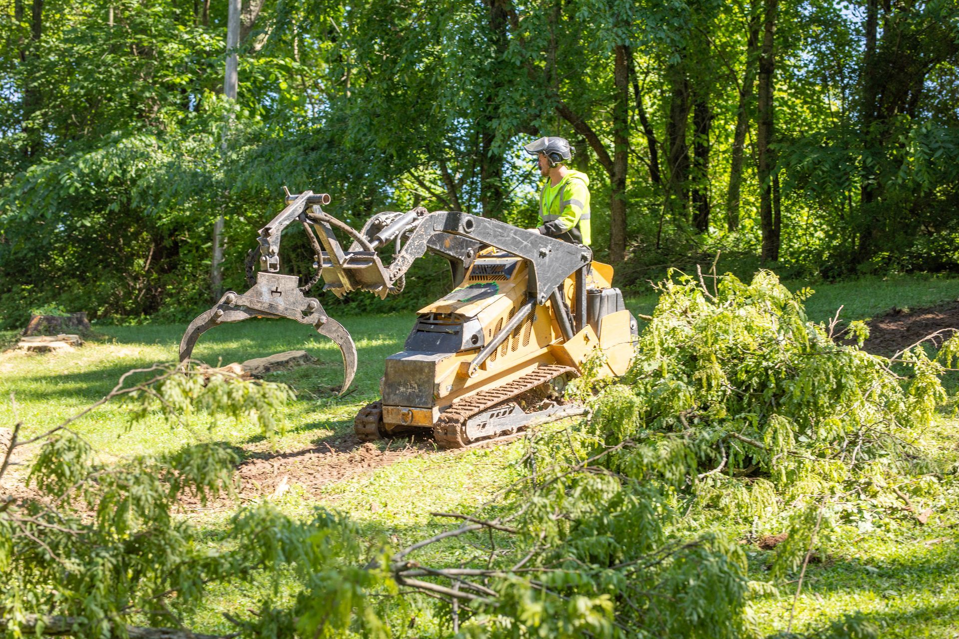 A yellow skid steer with a grapple attachment clearing branches and debris, operator in a wooded area.