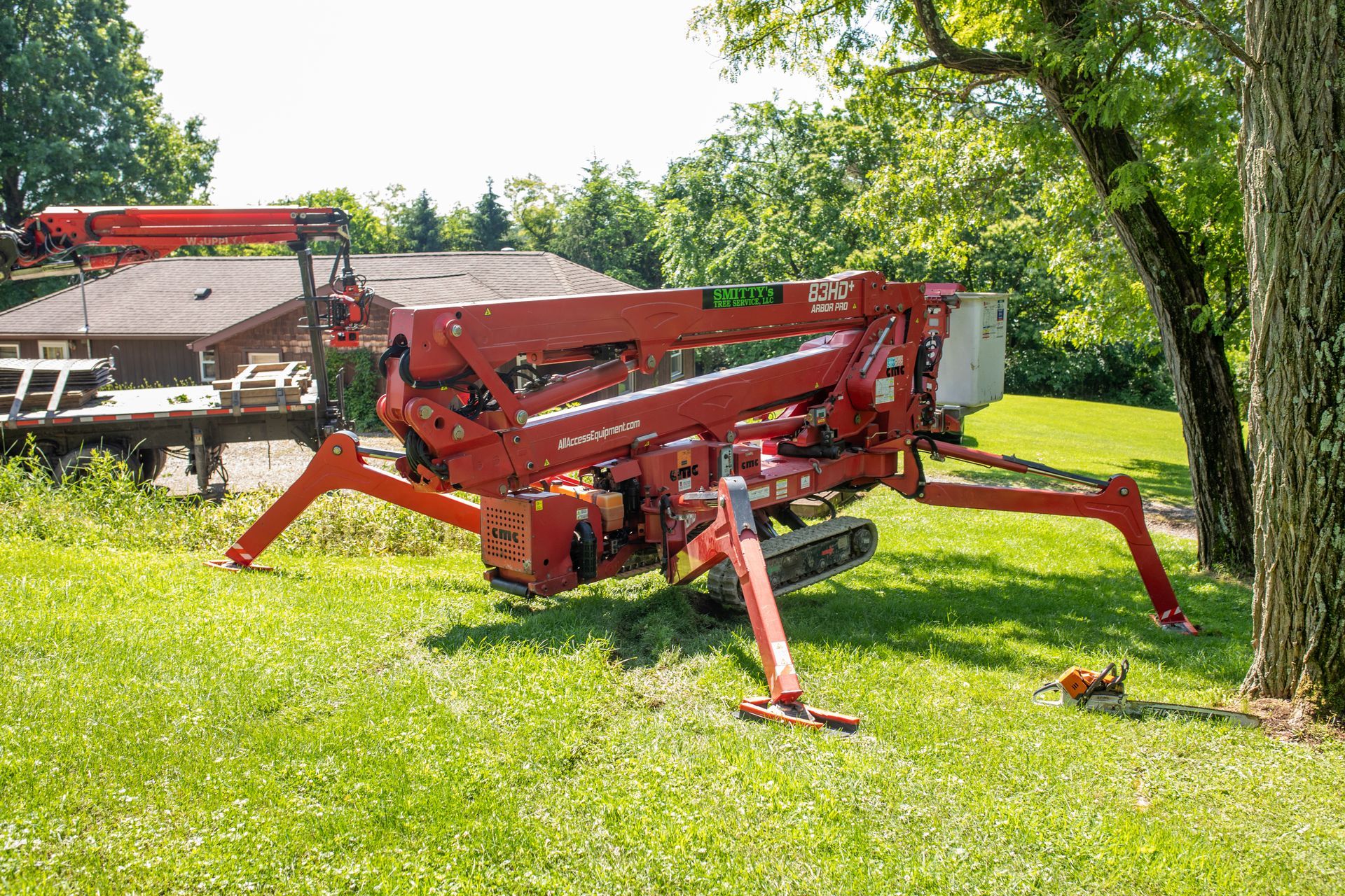 Red tree-trimming lift on a grassy lawn with outriggers extended, near a tree. A house and trailer are in the background.
