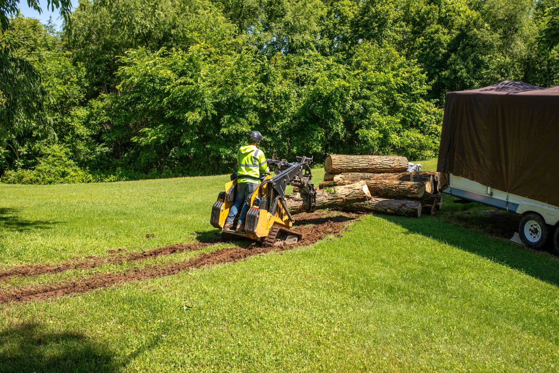 Person operating a small loader, moving logs onto a trailer in a grassy area near trees.