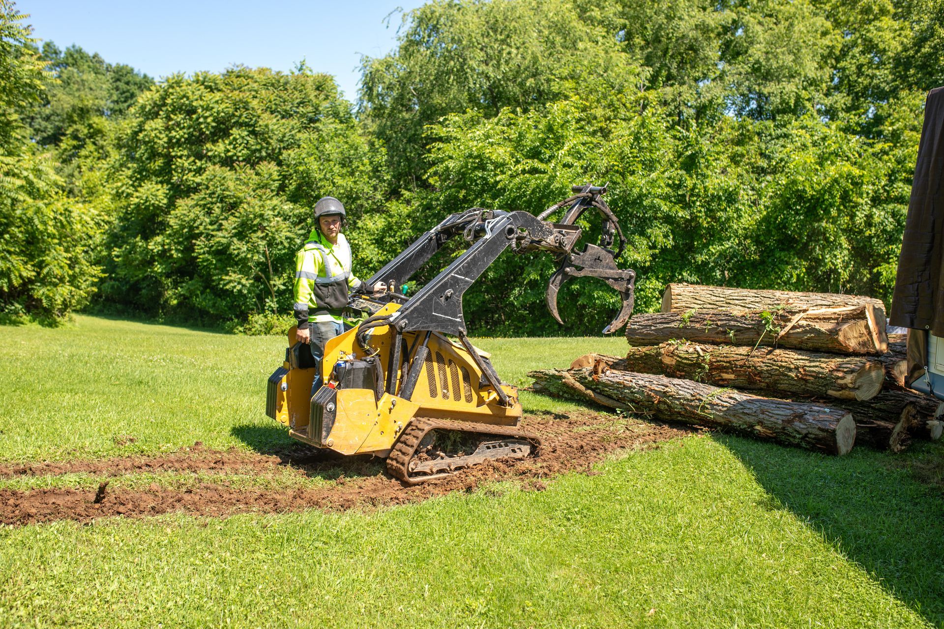 Person operating a yellow skid steer lifting logs in a grassy area with trees in the background.