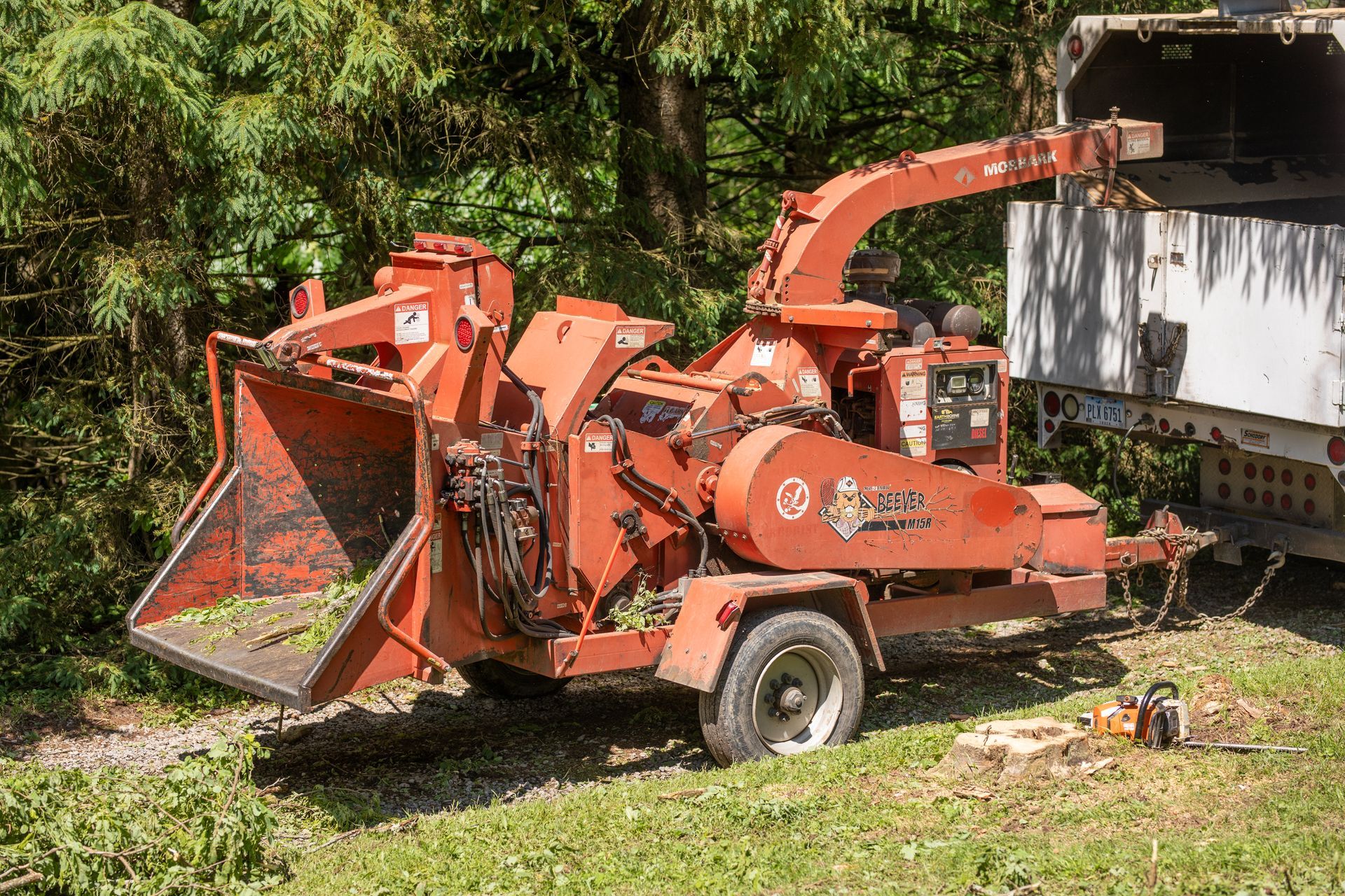 Red wood chipper next to a trailer, in a wooded area.