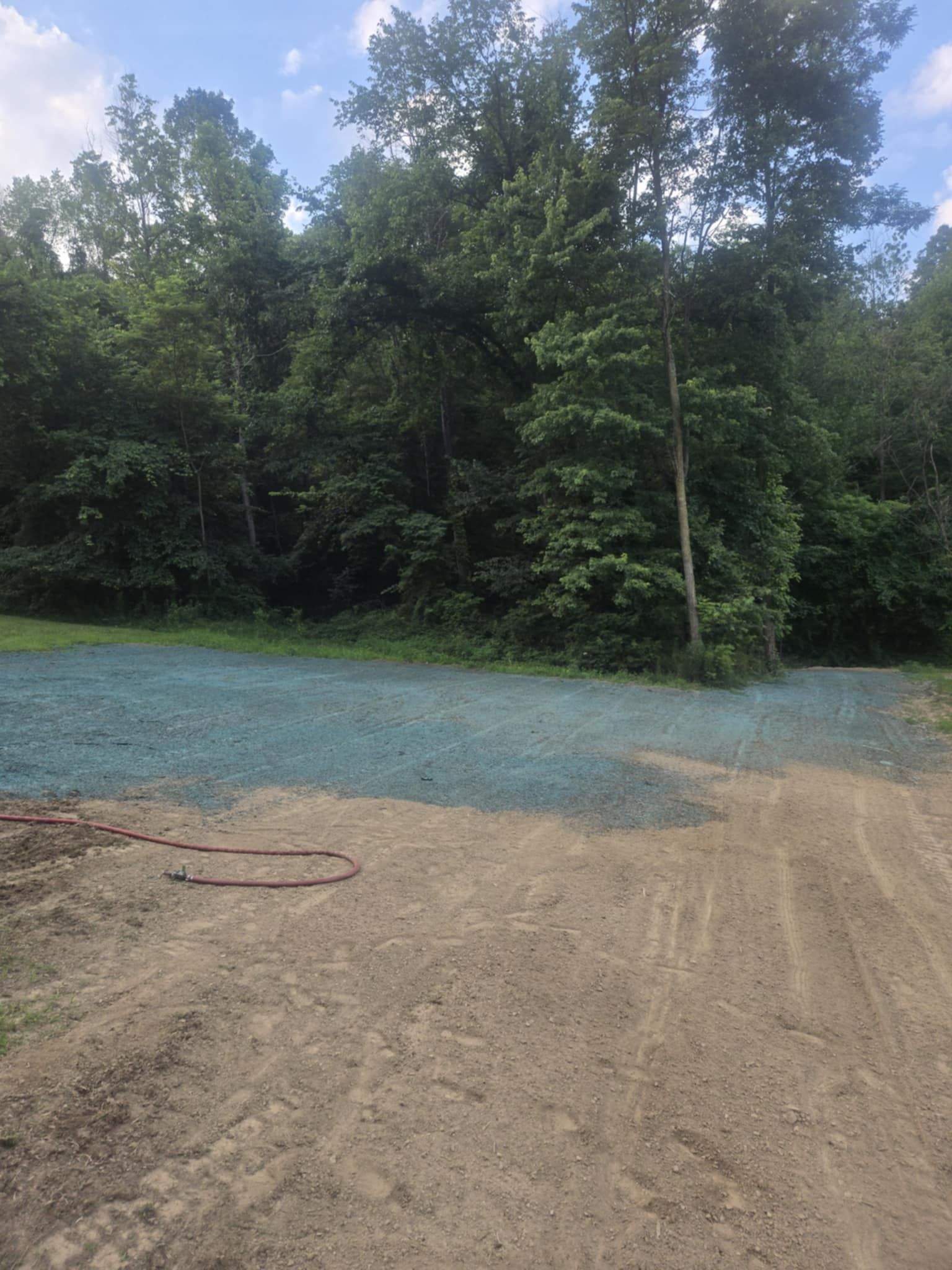 Dirt road transitions to blue-treated ground, leading to a tree line under a cloudy sky.