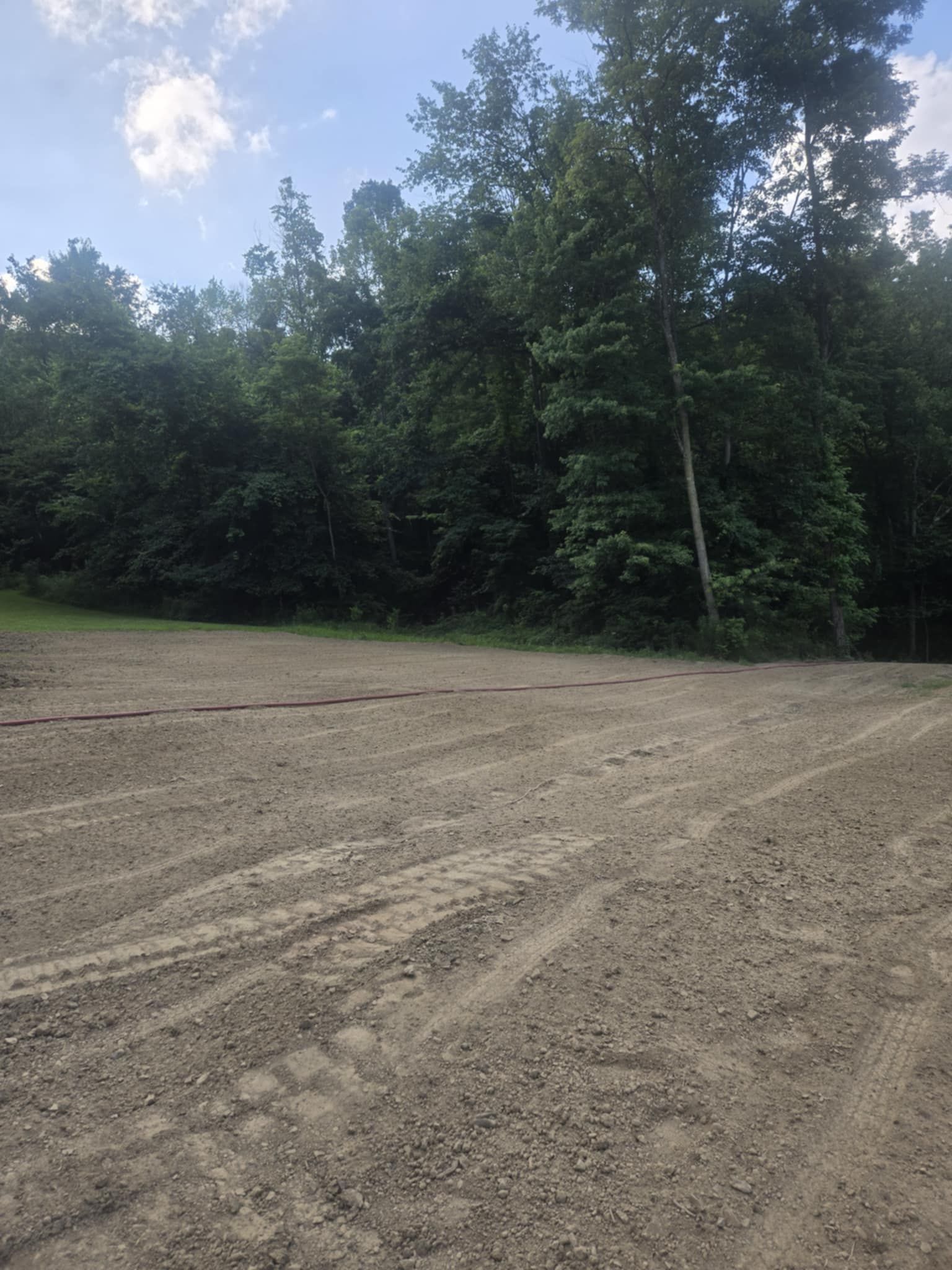 Gravel area with tire tracks in front of a line of trees under a cloudy sky.