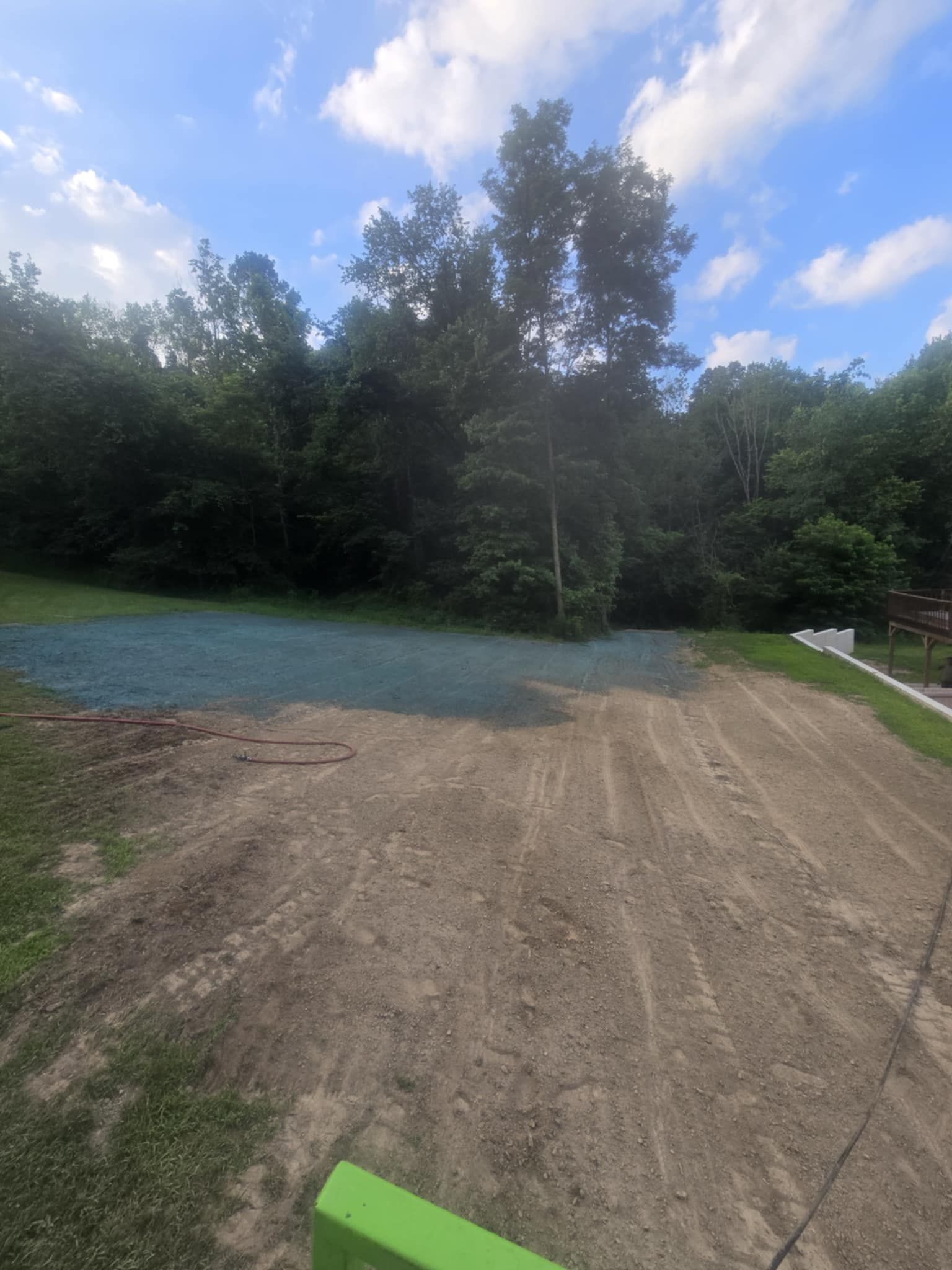 Cleared land, partially seeded with blue material, bordered by trees and grass, under a blue sky.