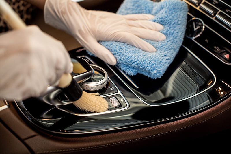 Person wearing gloves cleaning a car's interior with a brush and blue microfiber cloth.