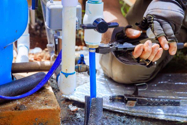 A pair of gloved hands uses pliers to secure a metal clamp on a blue PEX pipe connected to a white PVC plumbing valve.