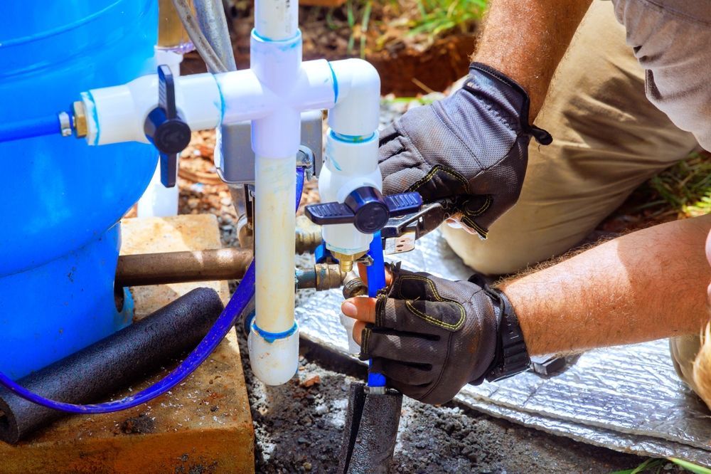 Gloved hands installing a blue PVC pipe valve beside a blue water barrel outdoors