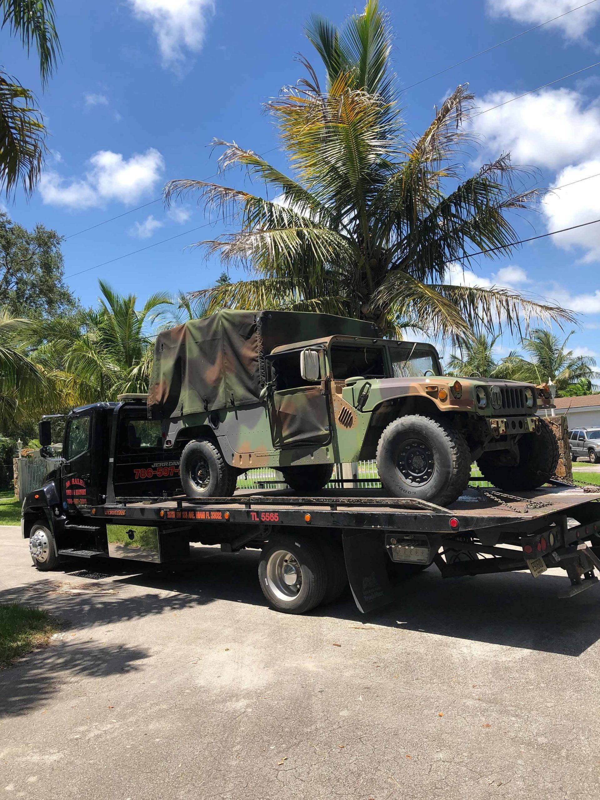 Military vehicle on a tow truck, camo, outdoors, sunny day.
