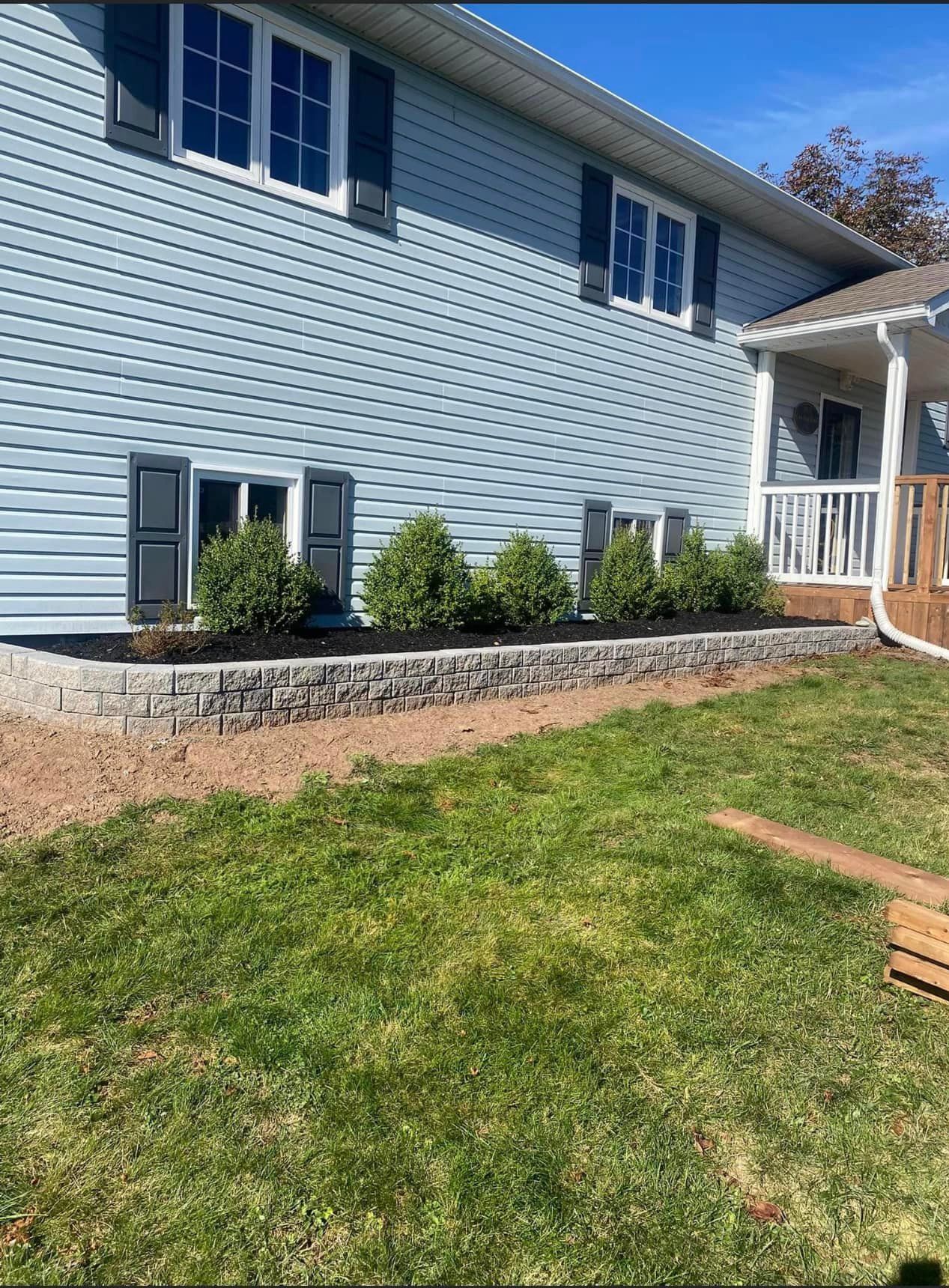 Light blue house exterior with shutters, stone planter bed, and green lawn under a sunny sky