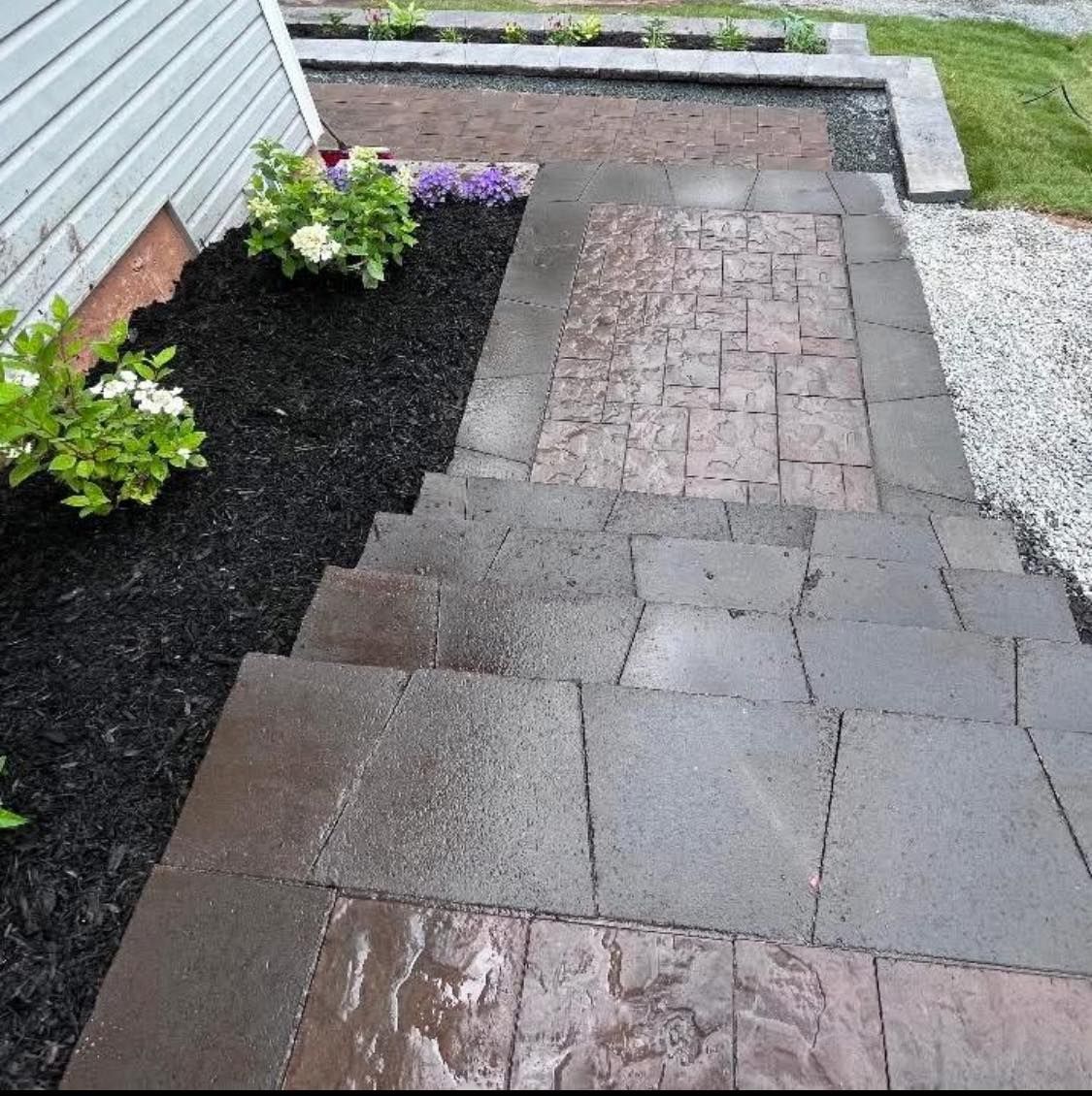 Wet brick and stone walkway beside a house, with black mulch flower bed and small flowers.