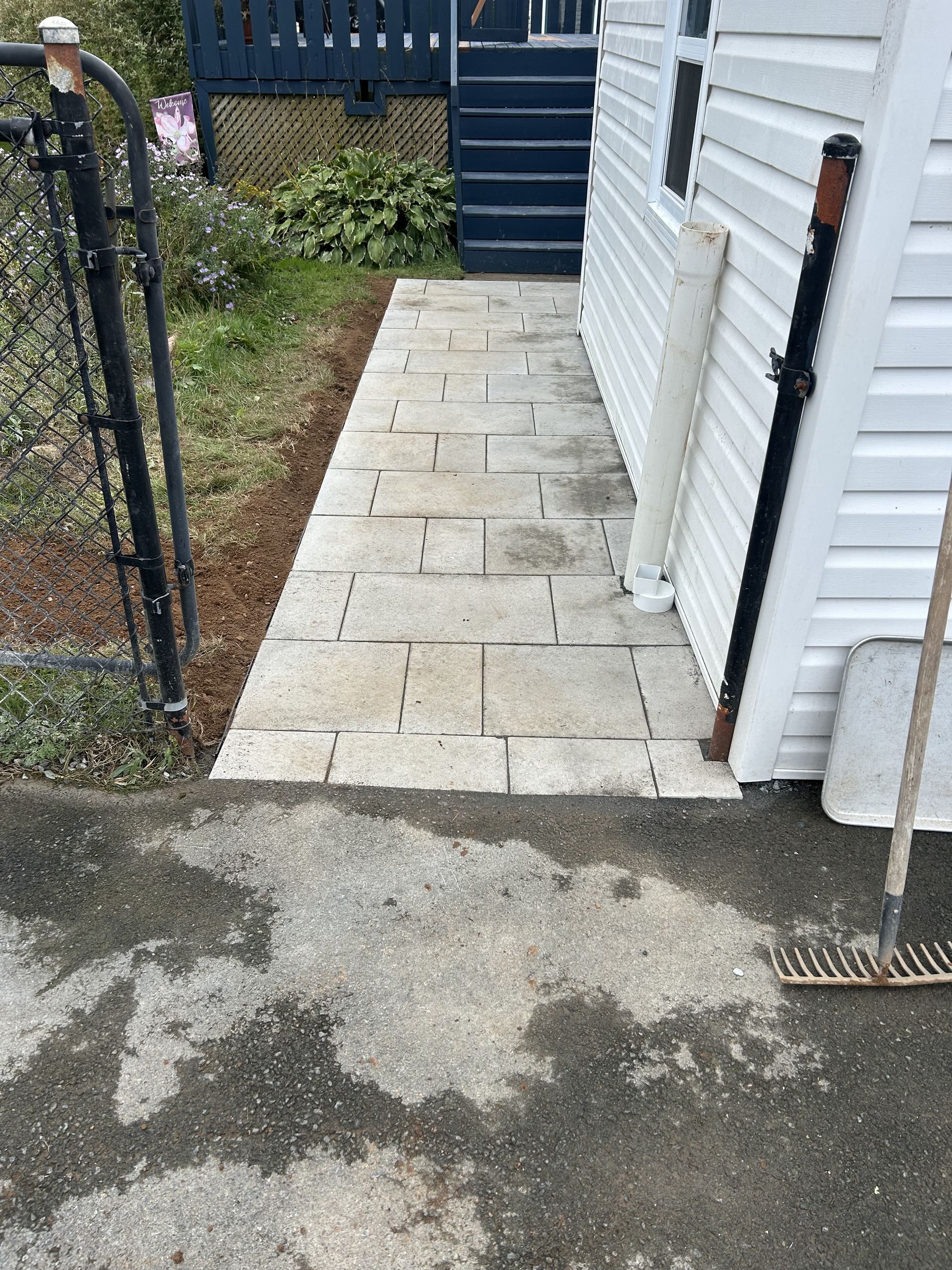 Narrow stone walkway beside a white building, with a black gate and patchy pavement in front