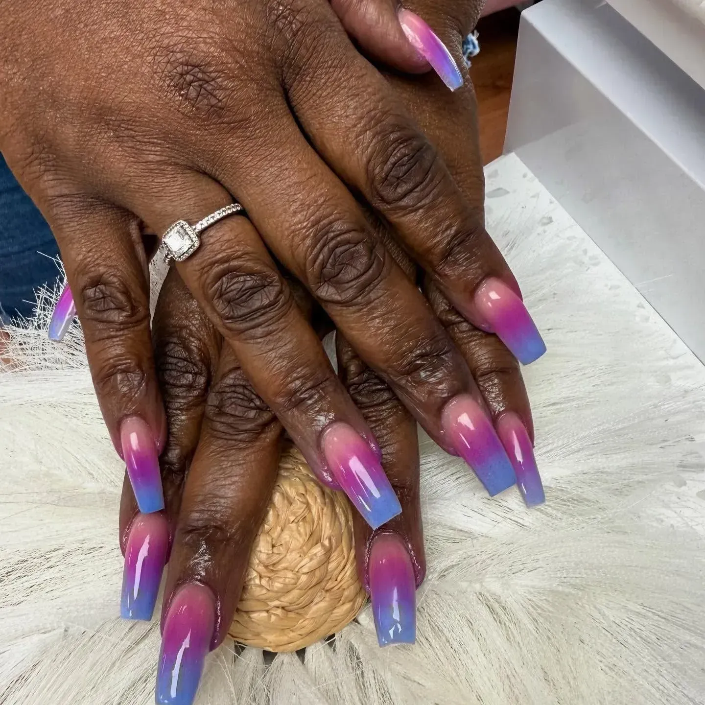 Hands with pink-to-blue ombré long nails resting on a textured beige surface.