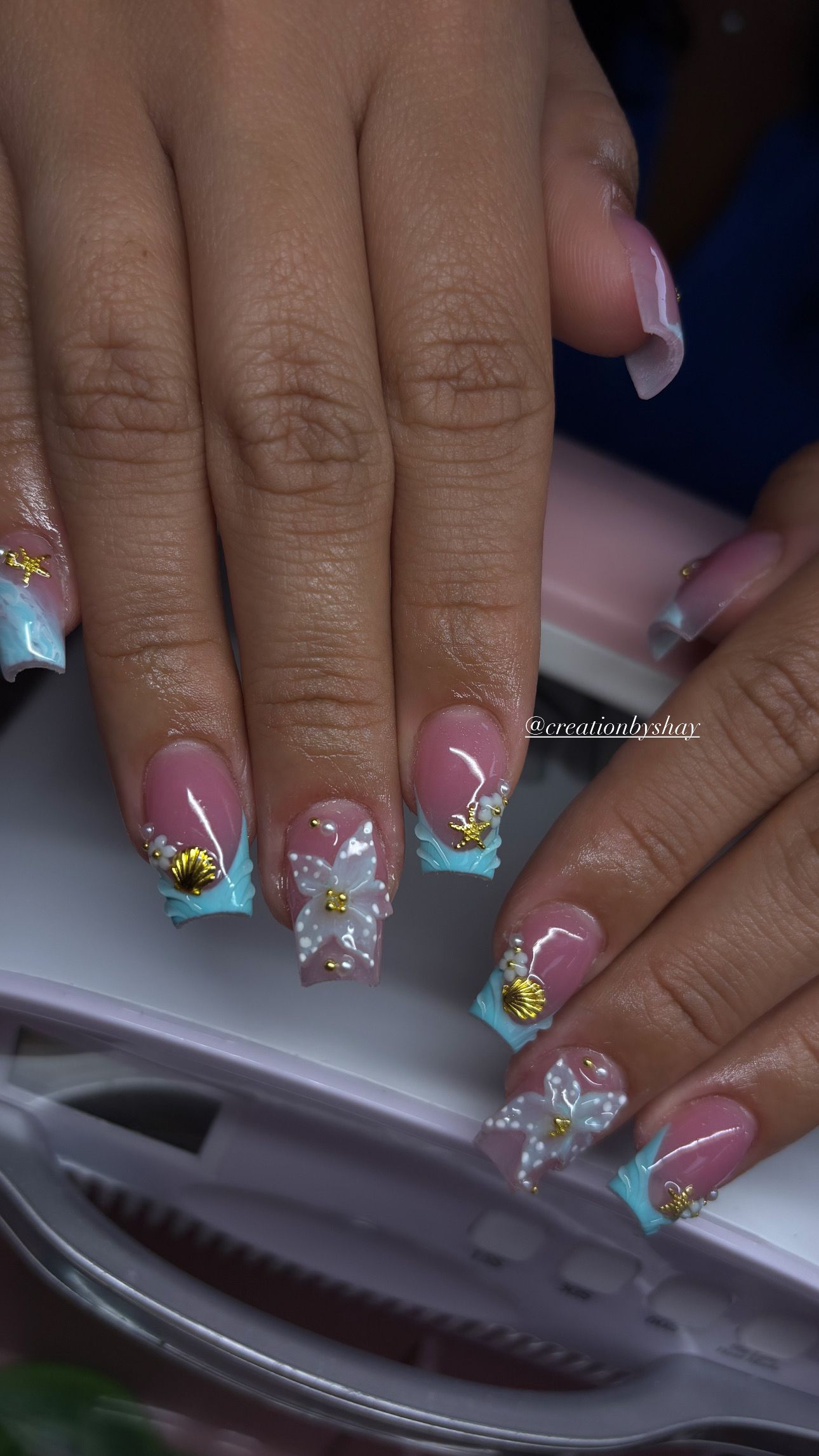 Close-up of hands with pink-and-blue glitter acrylic nails over a nail lamp.