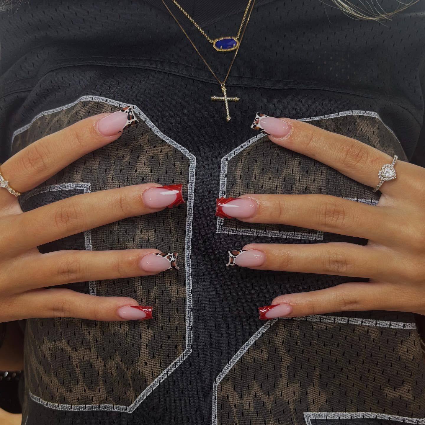 Hands with long pink nails and rings holding a sheer black patterned top, with layered necklaces visible.