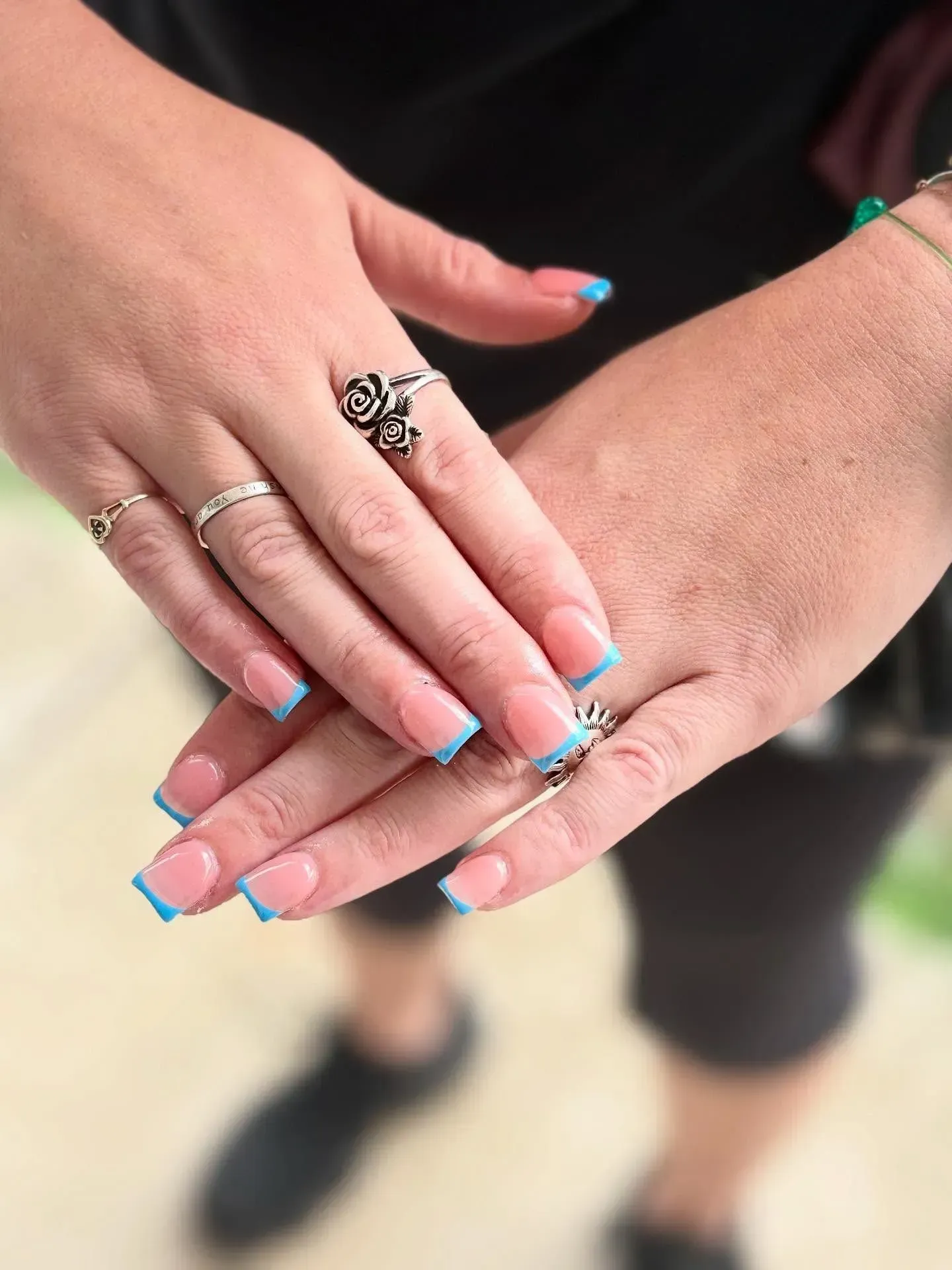 Hands with turquoise French manicure and rings, clasped together outdoors.